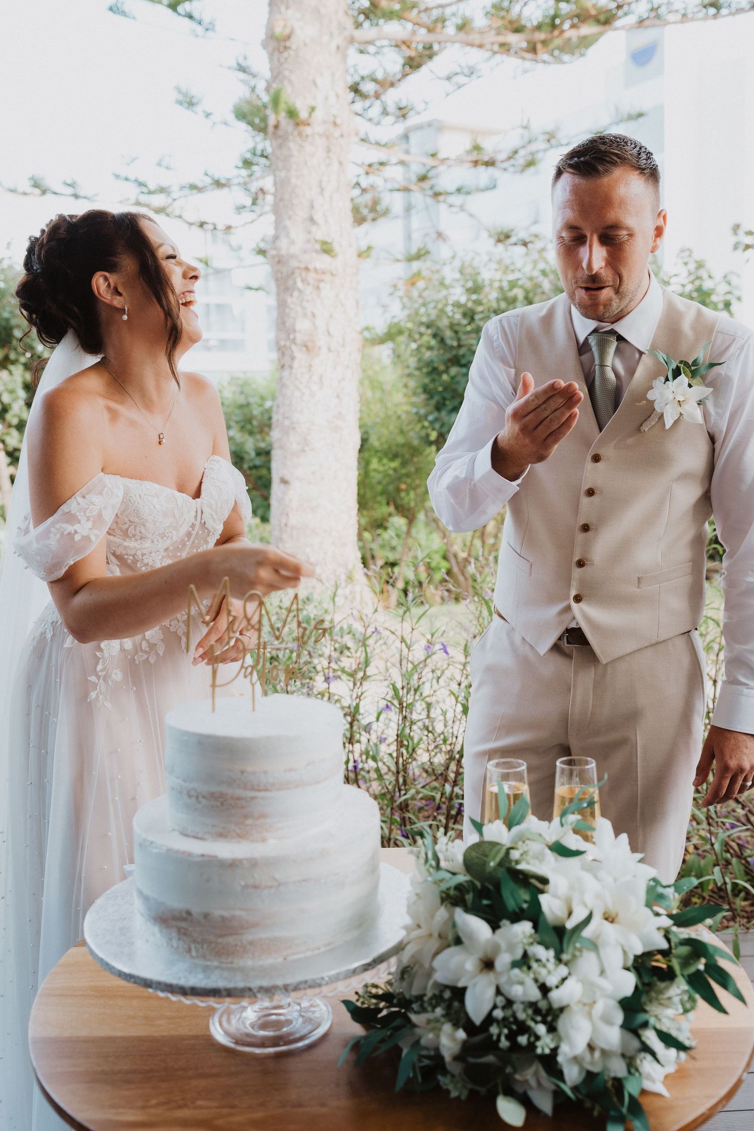 A couple laughs together at their wedding cake table decorated with white florals in an outdoor garden setting.
