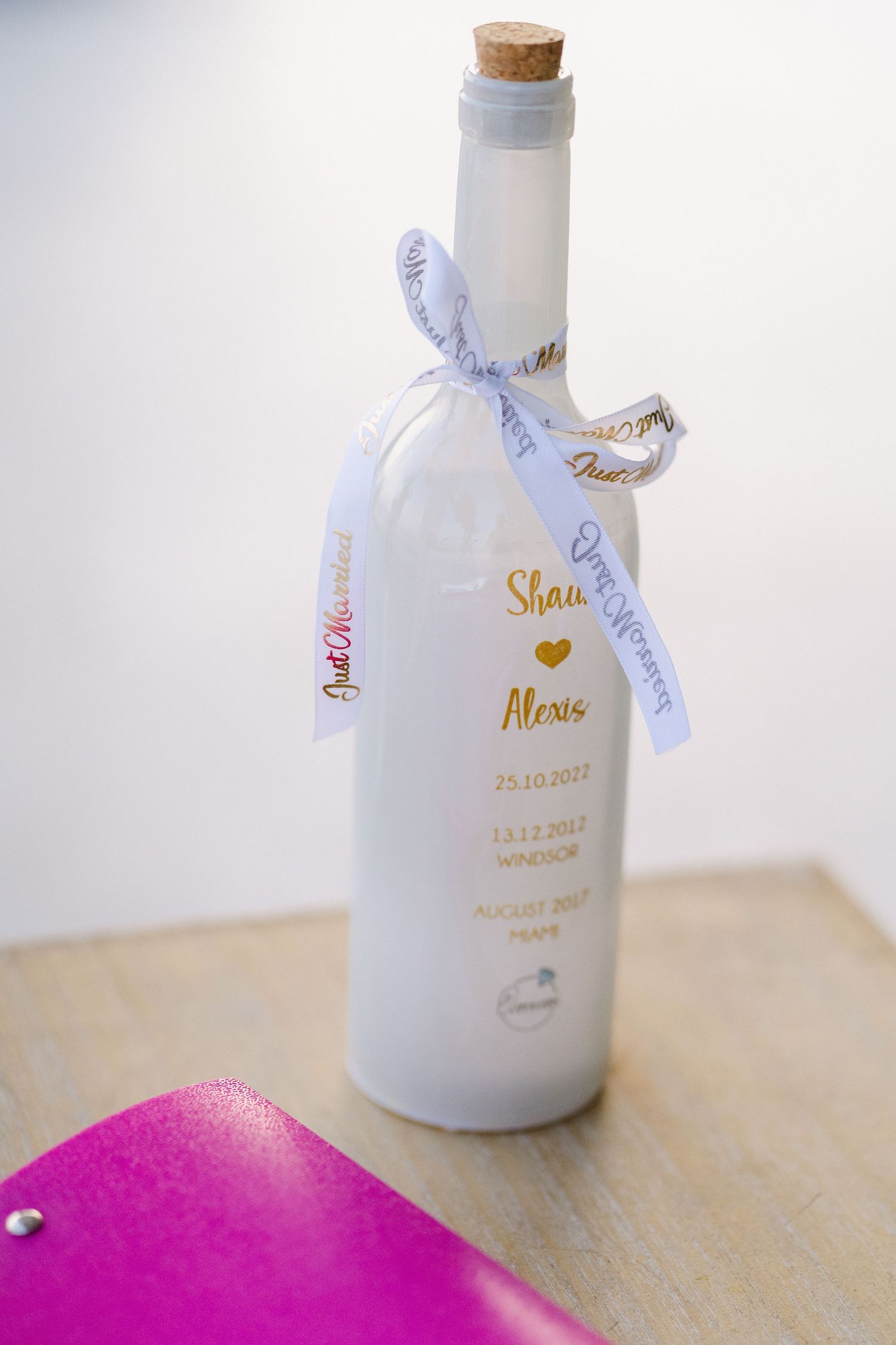 White frosted glass bottle with gold text and light blue ribbon as wedding favor.