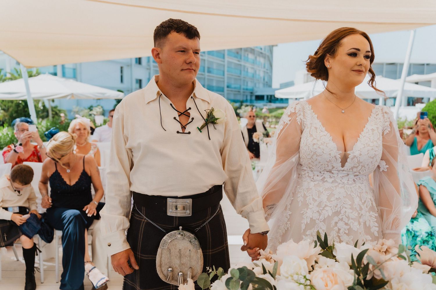 Couple in traditional Scottish and bridal attire holding hands during outdoor ceremony.