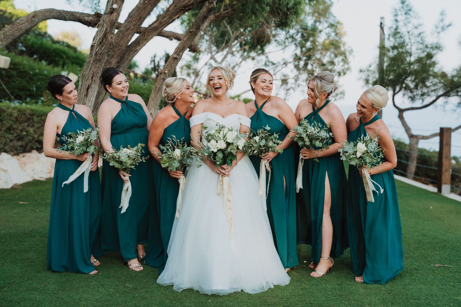 A bridal party poses outdoors wearing elegant deep teal bridesmaid dresses and holding coordinating floral bouquets.