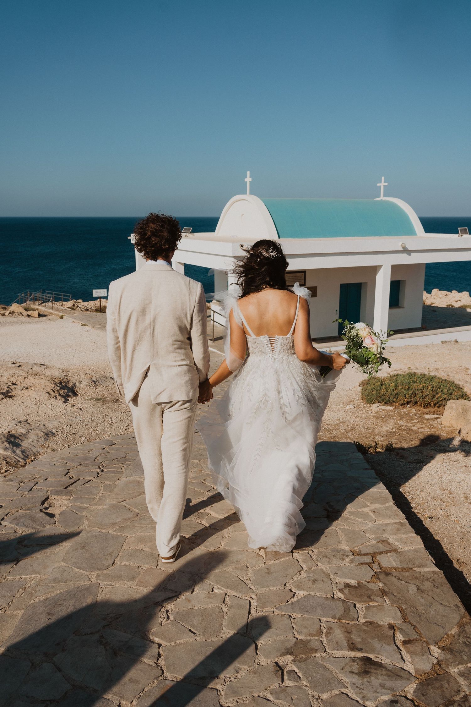 Couple walking towards white Mediterranean chapel overlooking blue ocean.