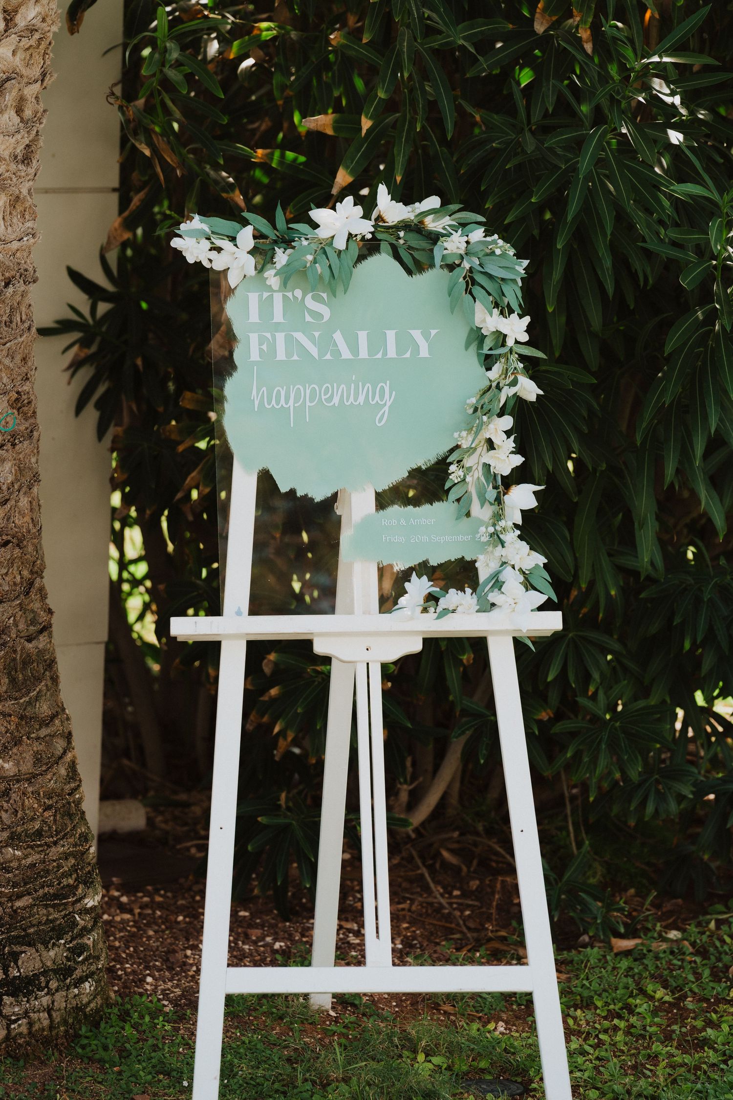 A white easel displays a mint green wedding welcome sign decorated with eucalyptus leaves and white flowers against foliage.