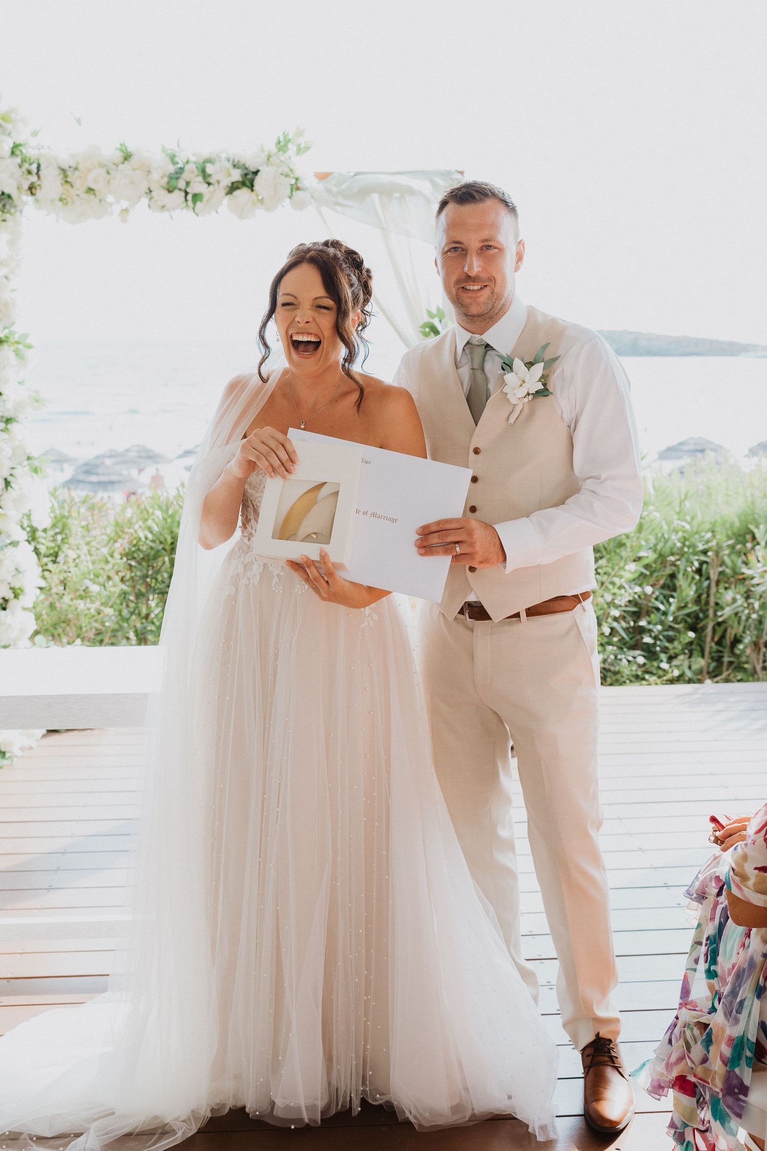 A joyful wedding couple sharing a laugh while holding paperwork during their outdoor ceremony with floral decor.