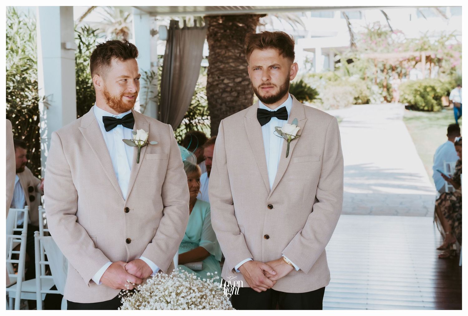 Groomsmen in beige suits with bow ties stand under covered outdoor ceremony space.