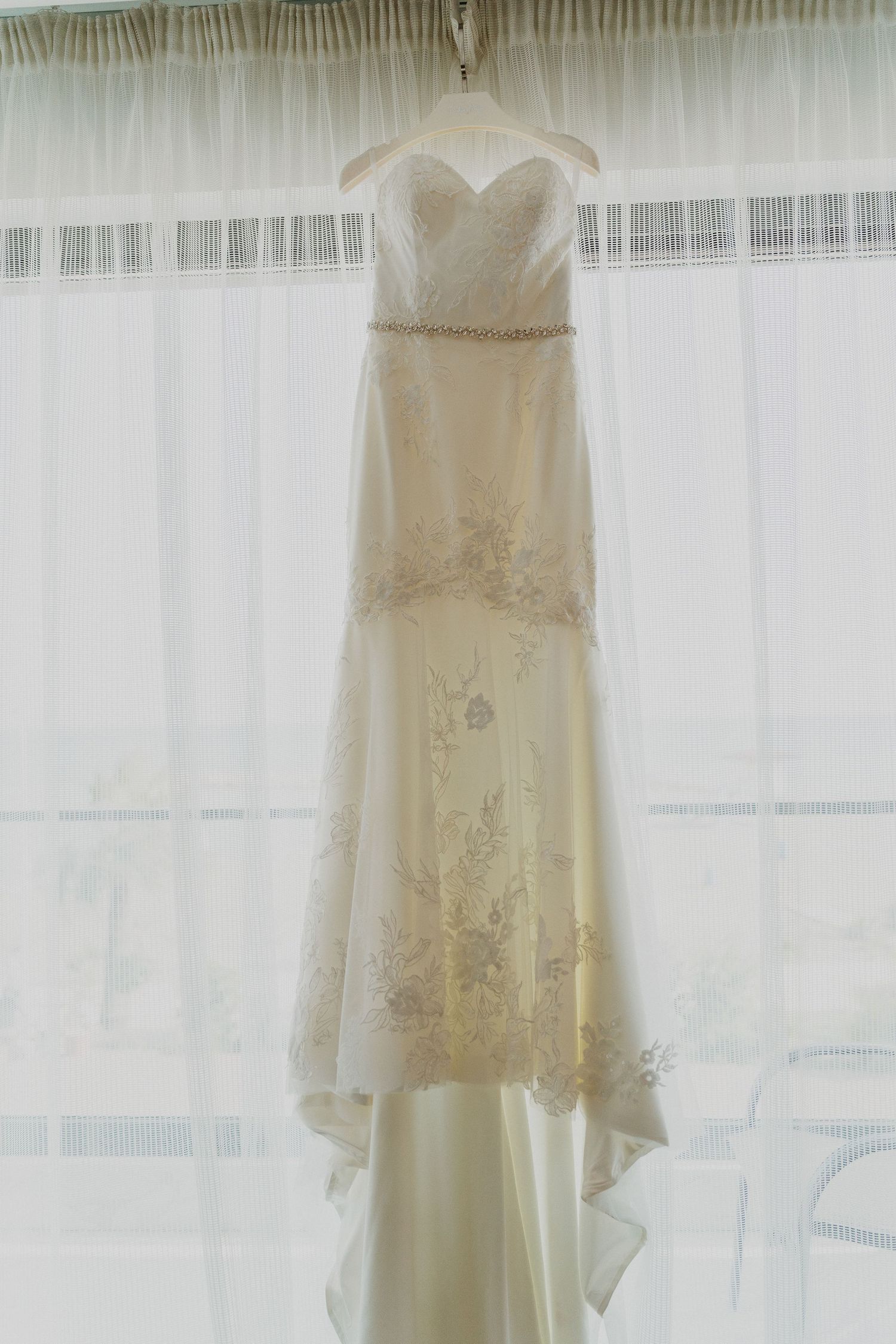 Elegant white wedding dress with lace details hanging in front of sheer curtains.