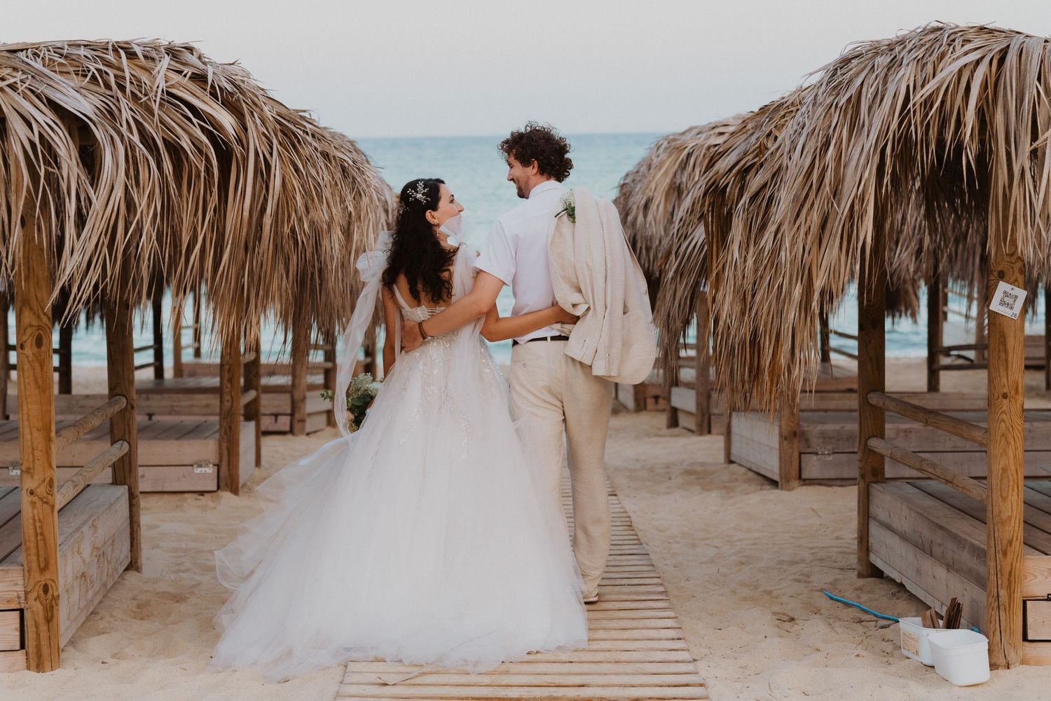 Beach wedding ceremony between rows of tiki umbrellas on wooden walkway.