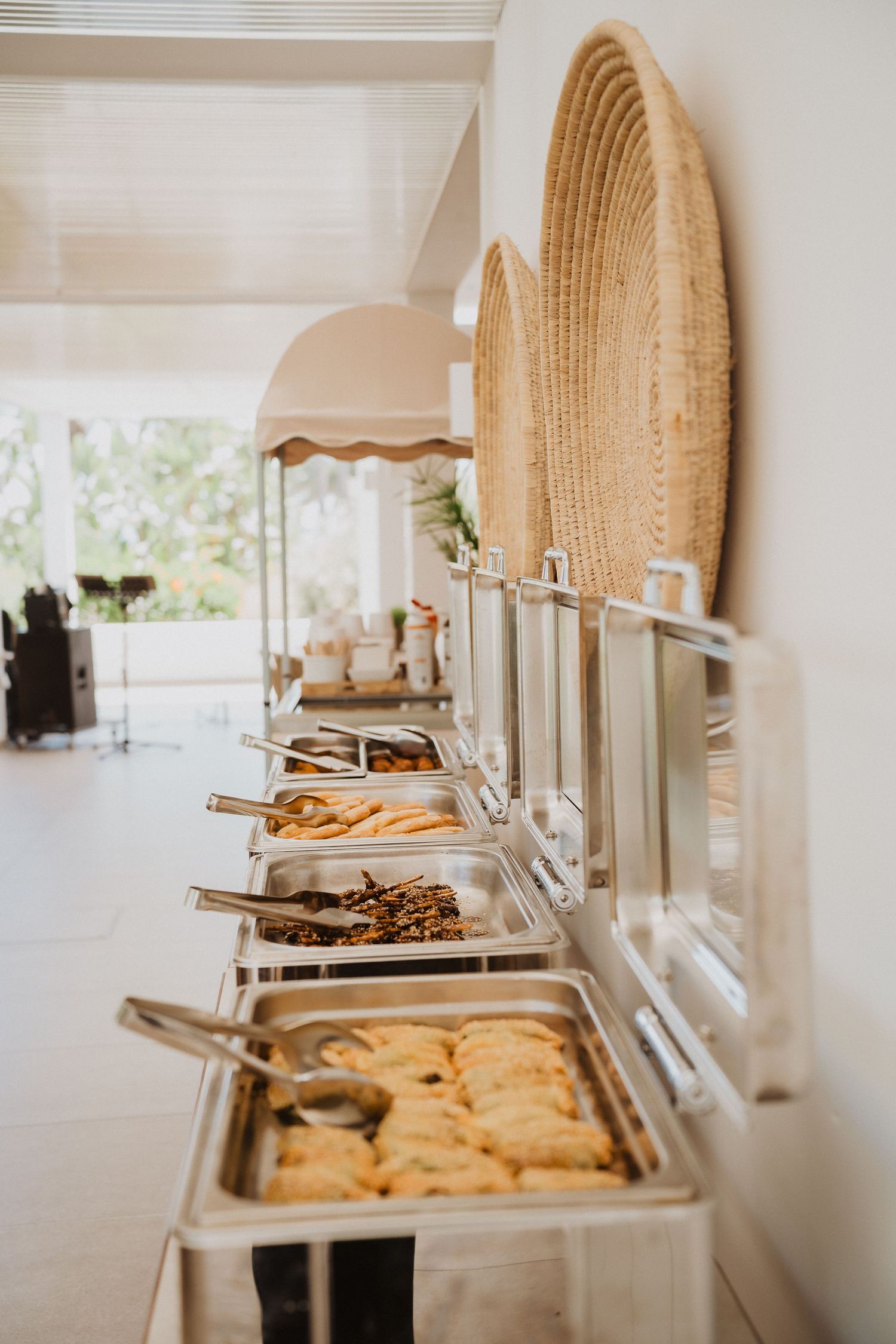 Elegant buffet setup with chafing dishes and woven decor on a white tablecloth at an outdoor venue.