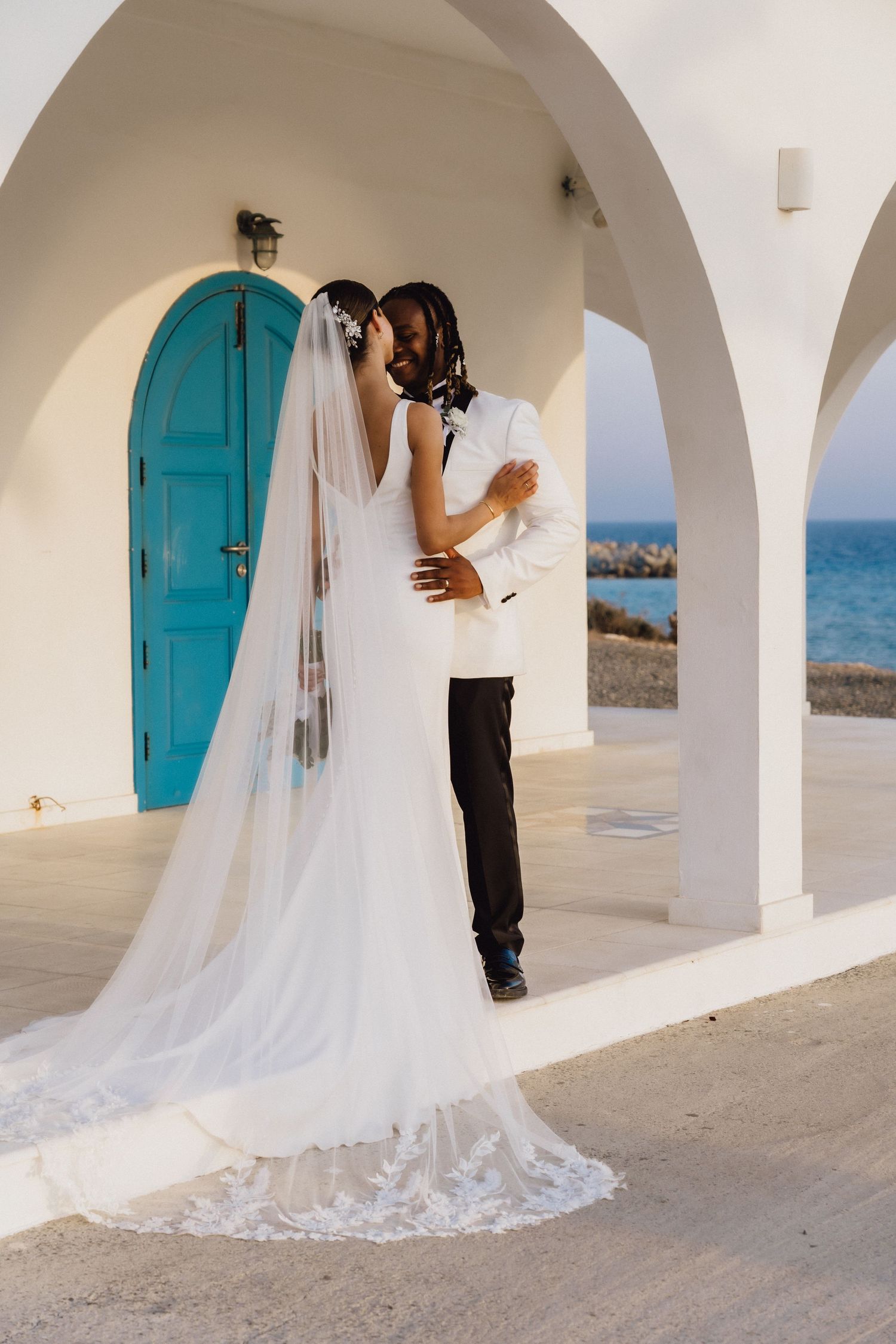 A romantic wedding moment captured under white Mediterranean arches with a turquoise door and ocean view.