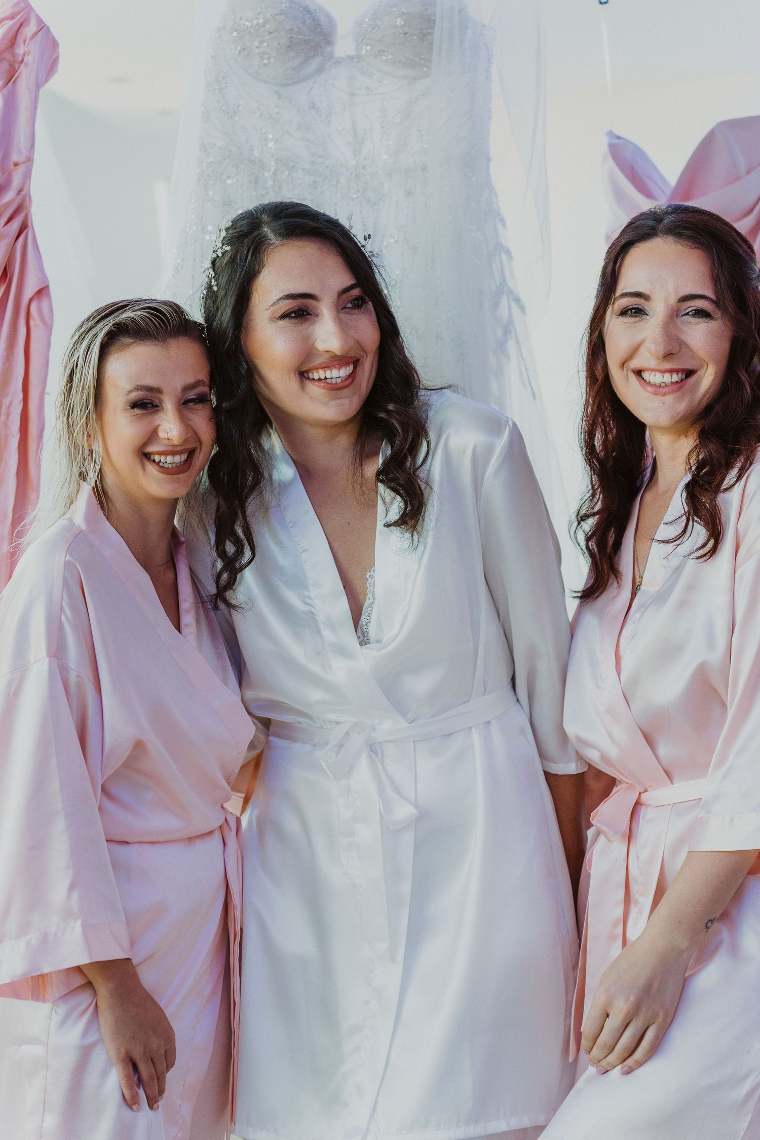 Three women in satin robes smile together during wedding preparations, with the bride in white and bridesmaids in pink.