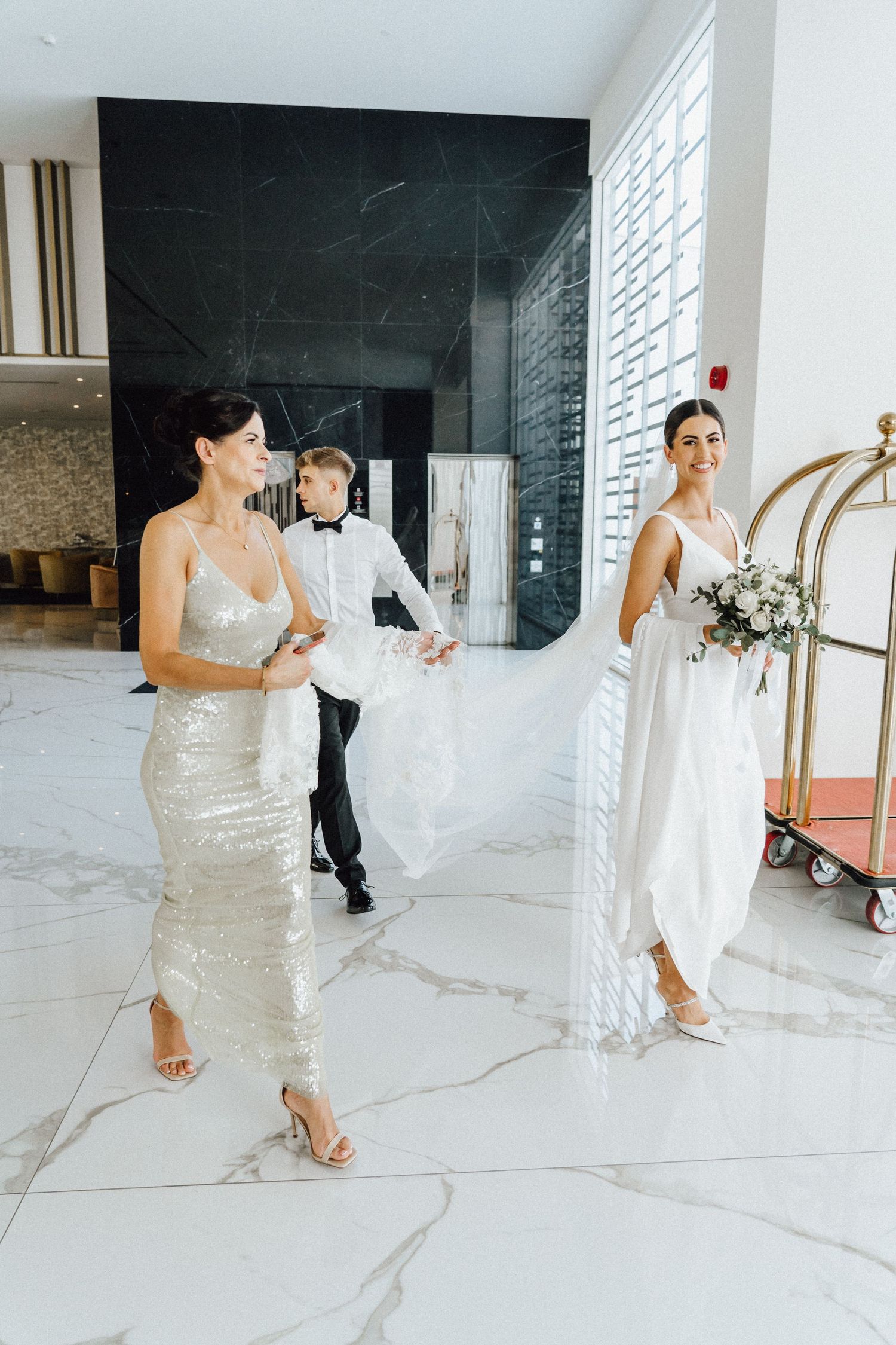 Wedding party walking through marble-floored lobby with luggage cart visible.