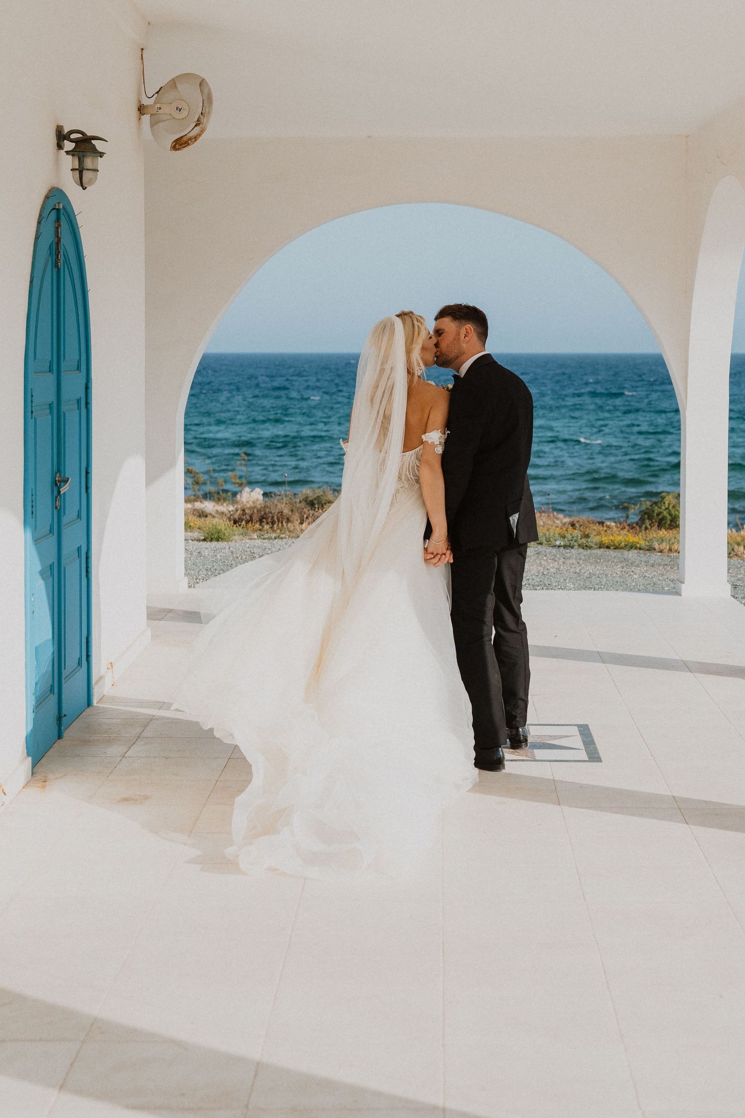 A coastal wedding portrait under white arches with turquoise doors and ocean views.