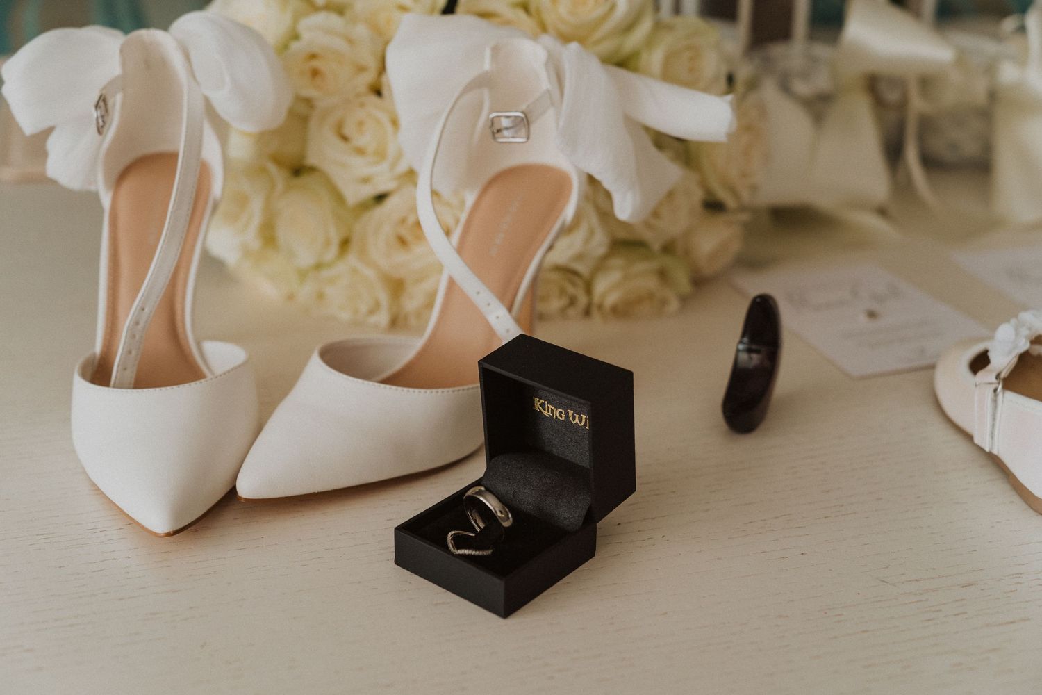White bridal heels and wedding rings in a black box arranged with white roses on a light surface.