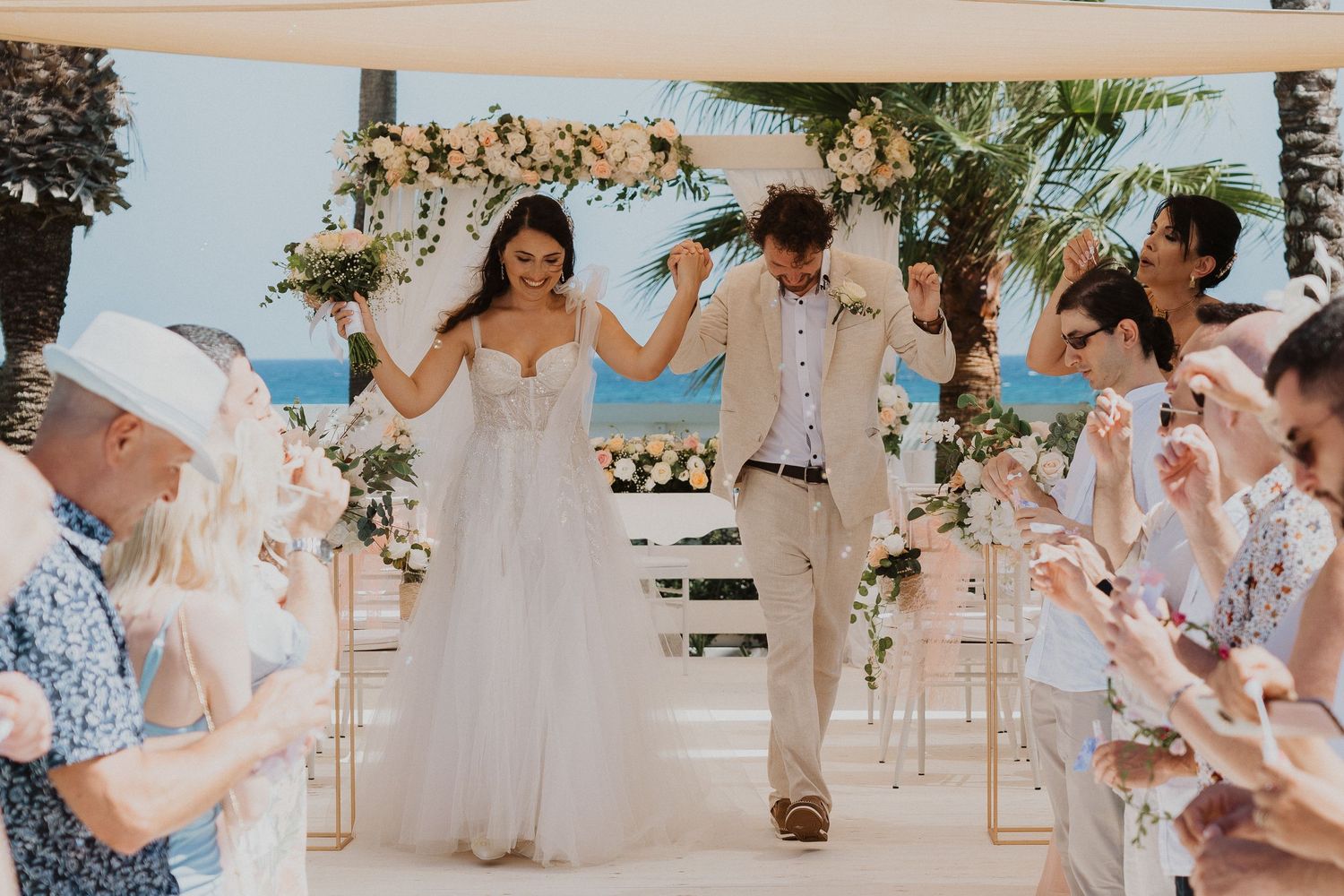 Wedding couple exits ceremony under flower arch while guests celebrate with bubble wands.