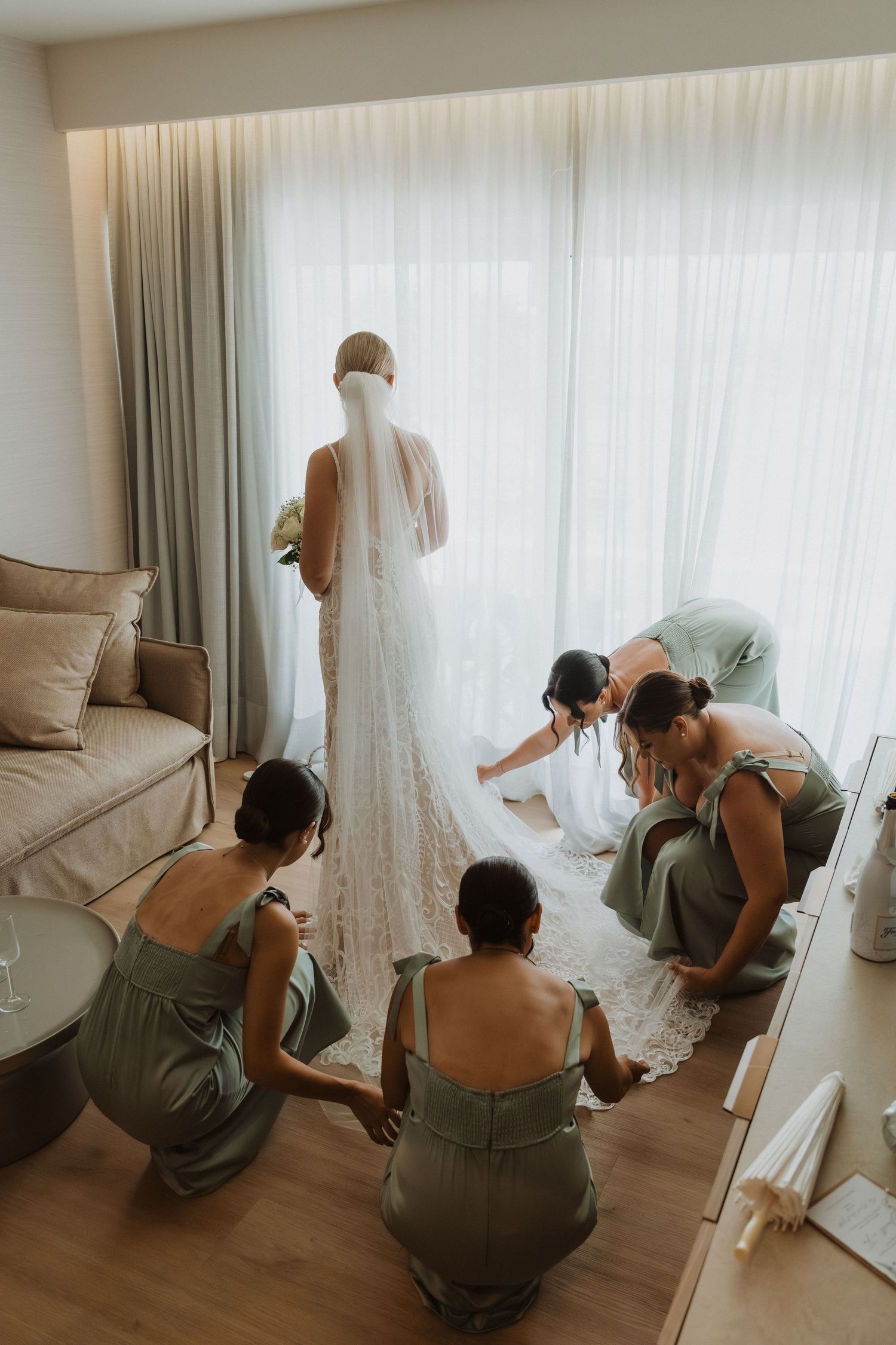 Wedding party helps with final dress preparations in a sunlit room with sage green bridesmaid dresses.