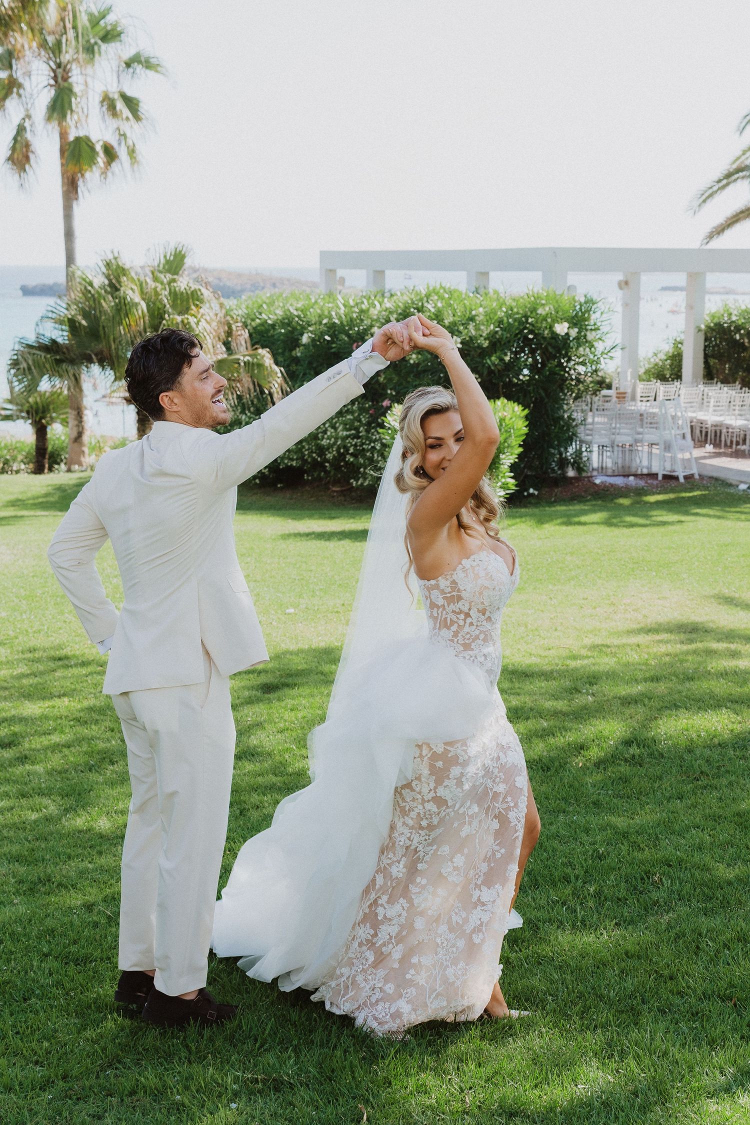 A couple dances playfully on a sunny lawn with palm trees and greenery in the background during a wedding celebration.