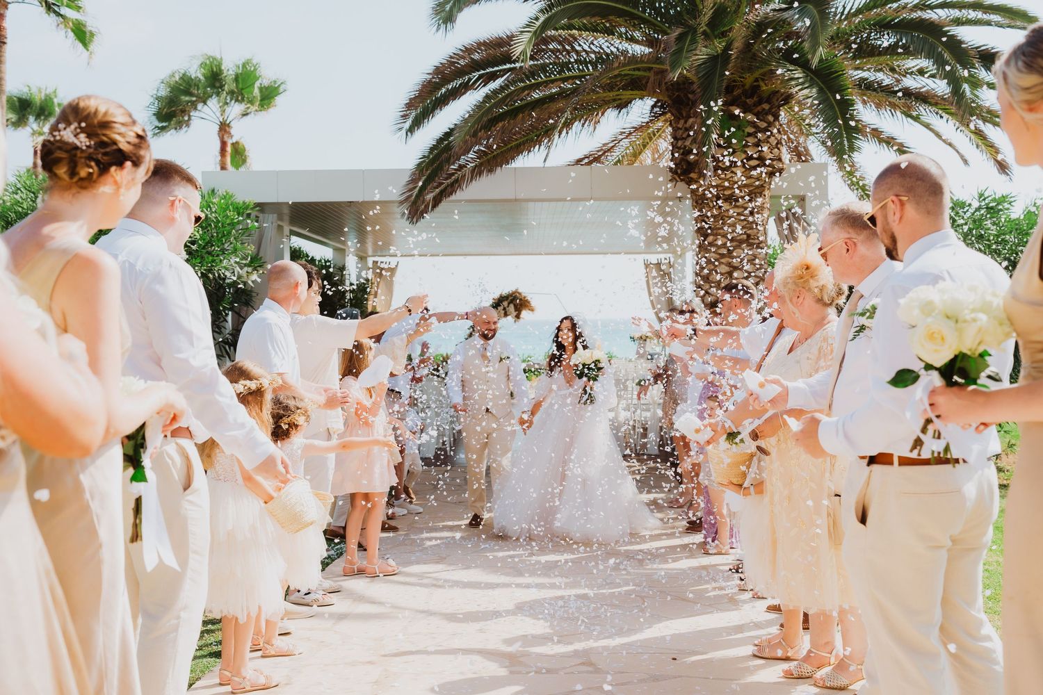 Wedding guests throw confetti in celebration under palm trees at a sunny outdoor ceremony.