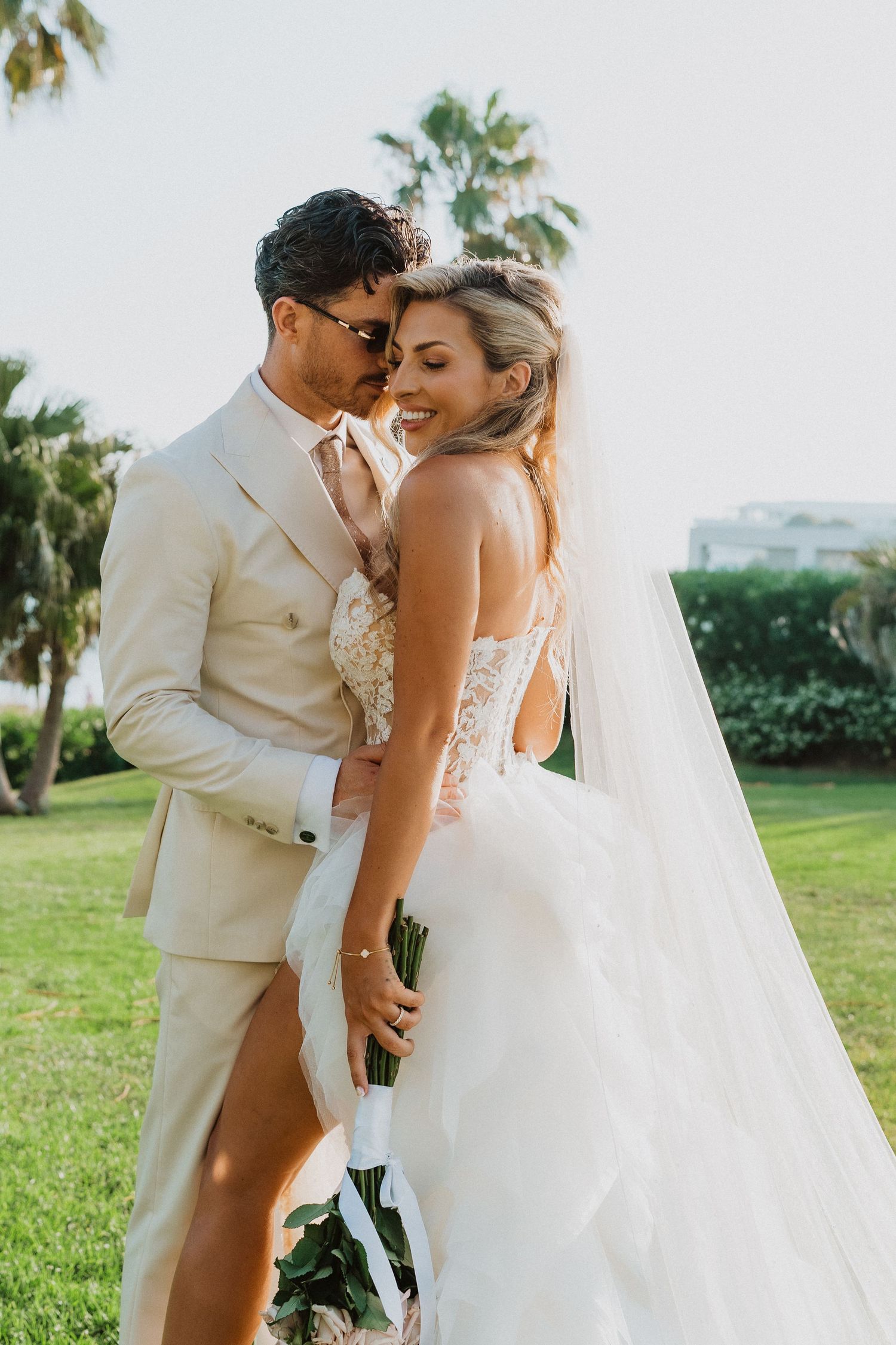 Romantic wedding couple embraces outdoors on a sunny day with palm trees and greenery in the background.