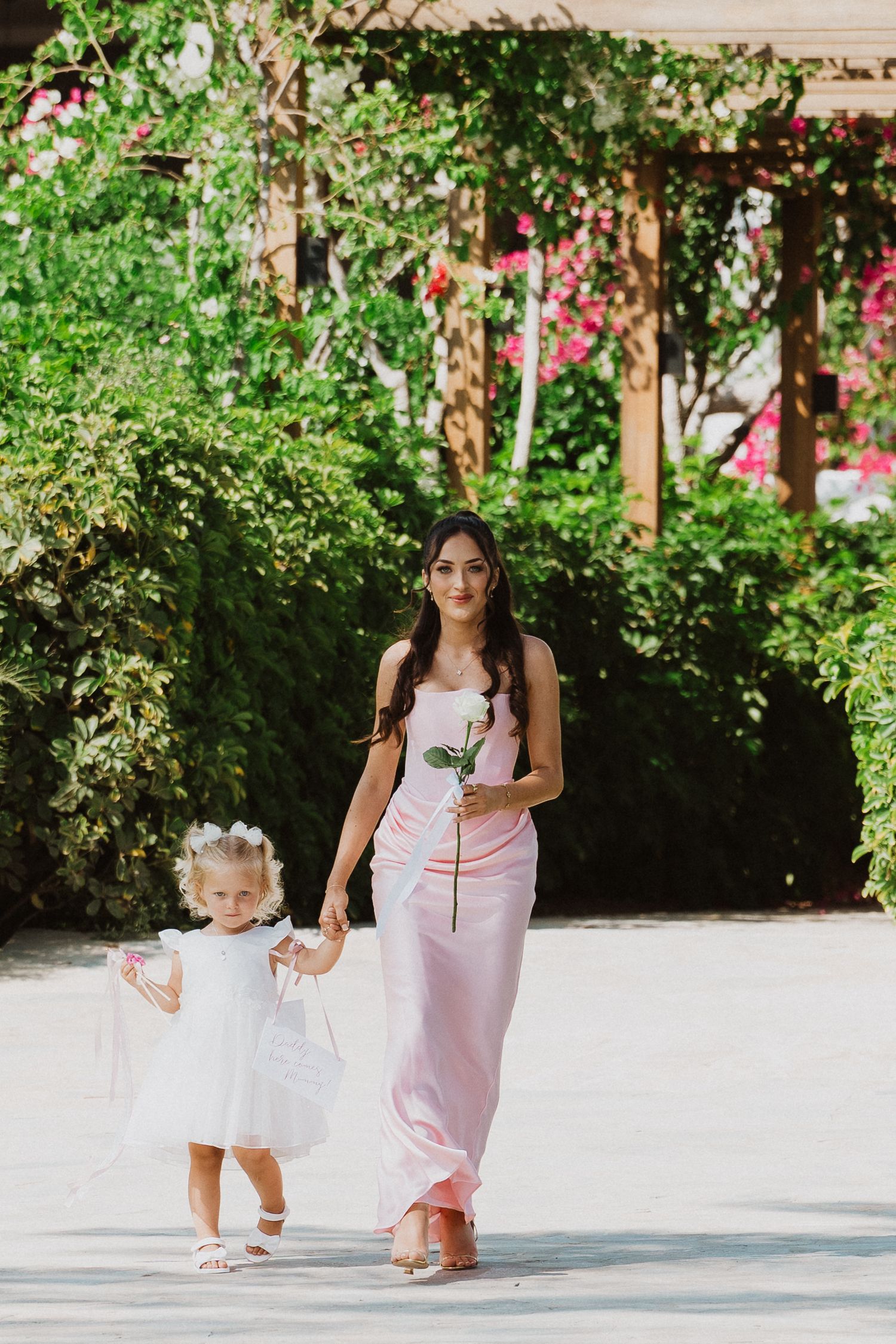 A pair in pink and white dresses walk hand in hand through a lush garden setting on a sunny day.