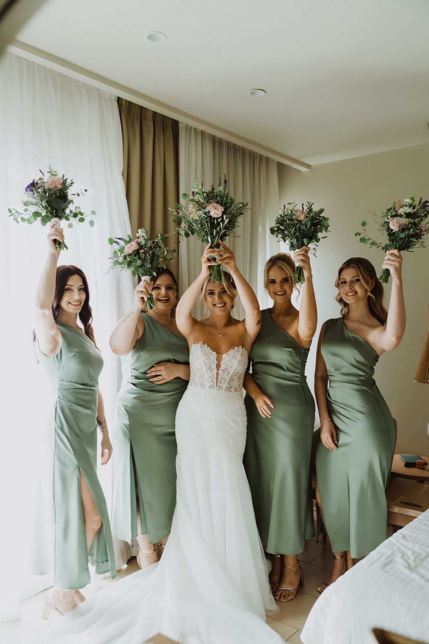 Candid moment of bridal party celebrating in sage green dresses by window light.