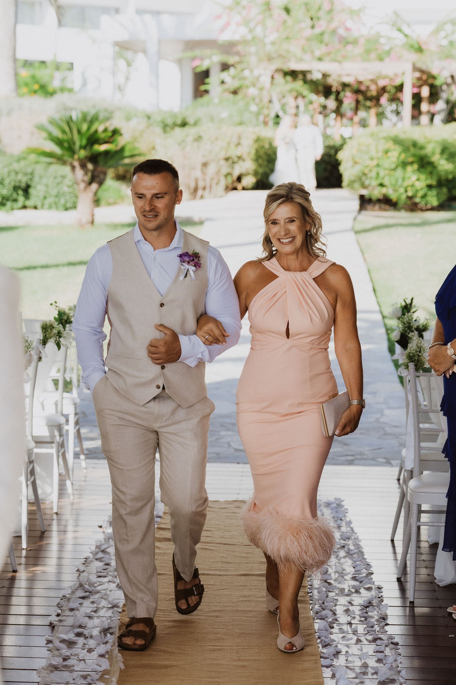 Wedding party members in beige and peach attire walk down an aisle lined with white petals.