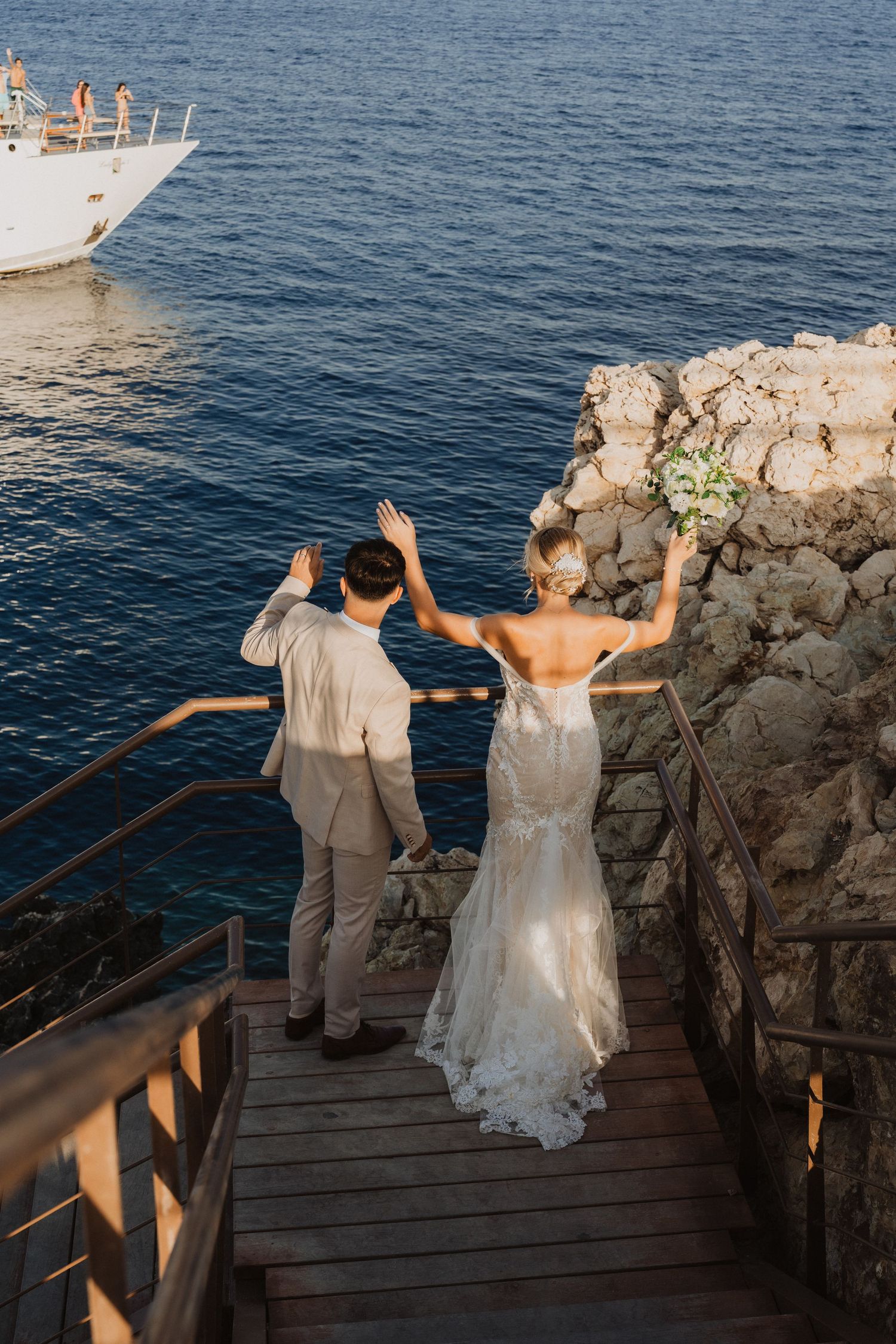 Wedding couple waves to passing boat while standing on coastal boardwalk.
