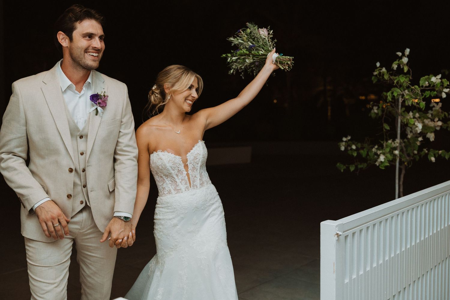 A joyful wedding couple walking together at night with the bride holding her bouquet high.