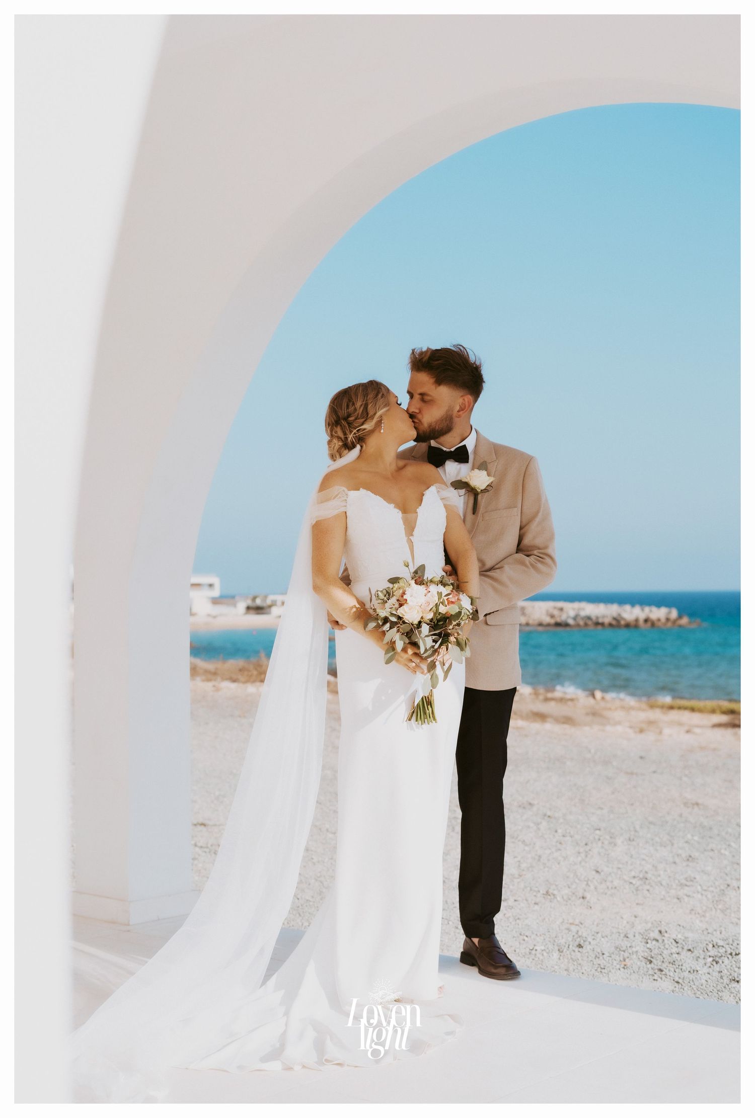 Romantic wedding portrait framed by white Mediterranean arch overlooking blue ocean waters.
