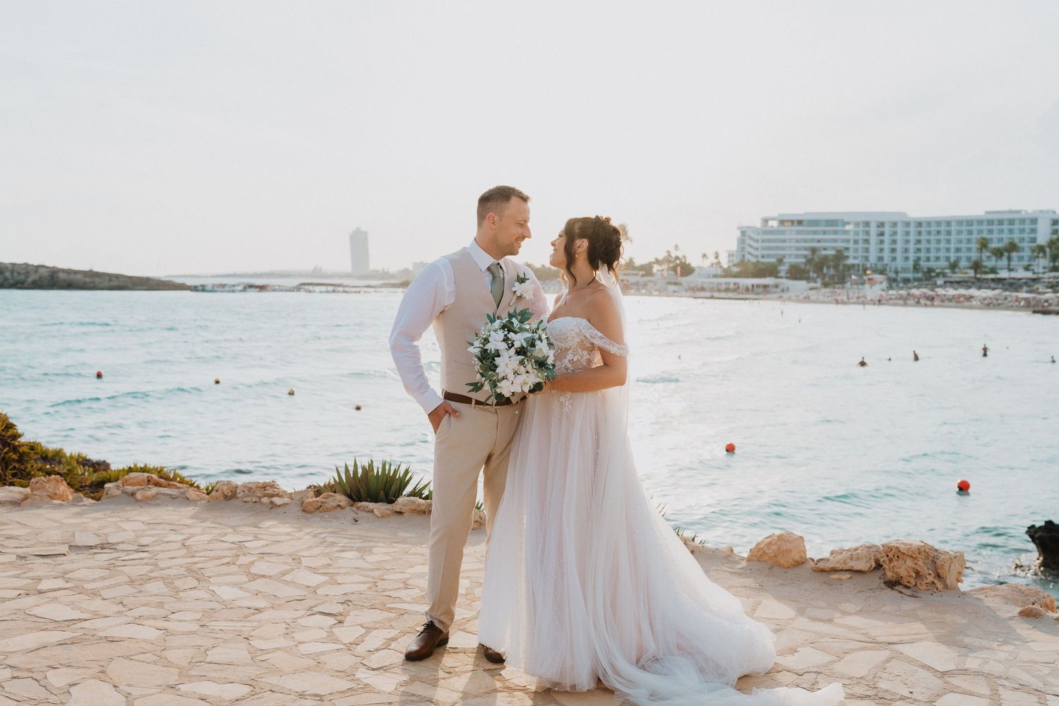 A wedding couple stands on a rocky coastal path overlooking a beach and resort buildings in the distance.