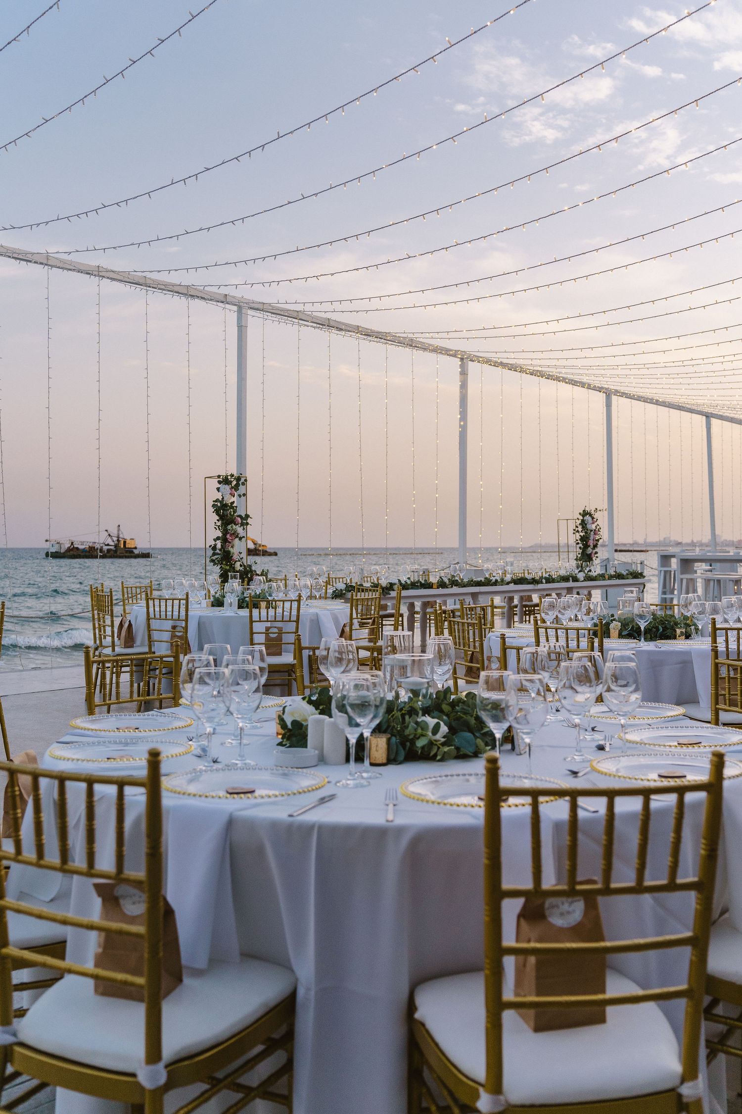 Seaside wedding reception tables with gold chairs and string lights creating a romantic atmosphere.