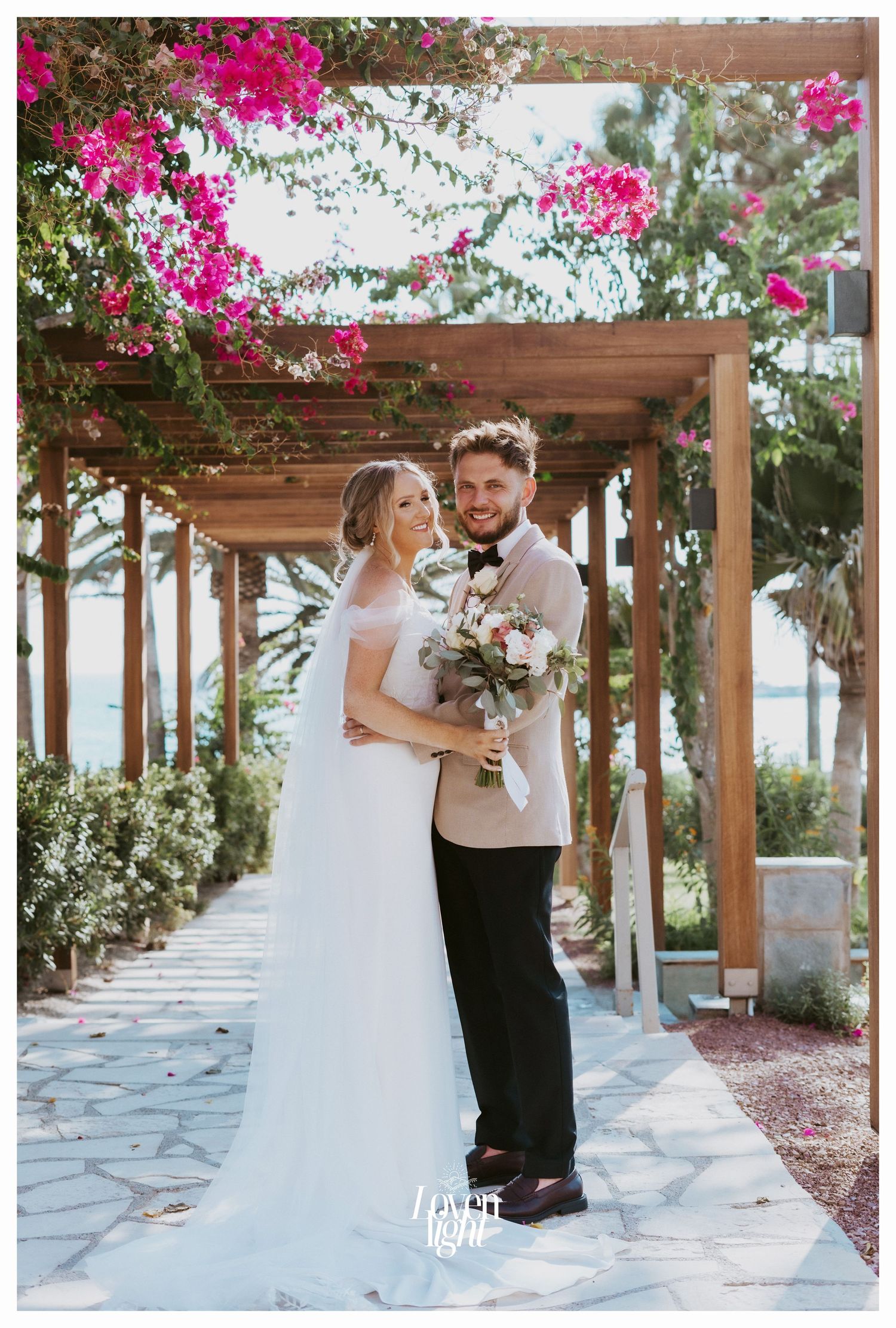 A newlywed couple poses under a wooden pergola decorated with vibrant pink bougainvillea flowers.