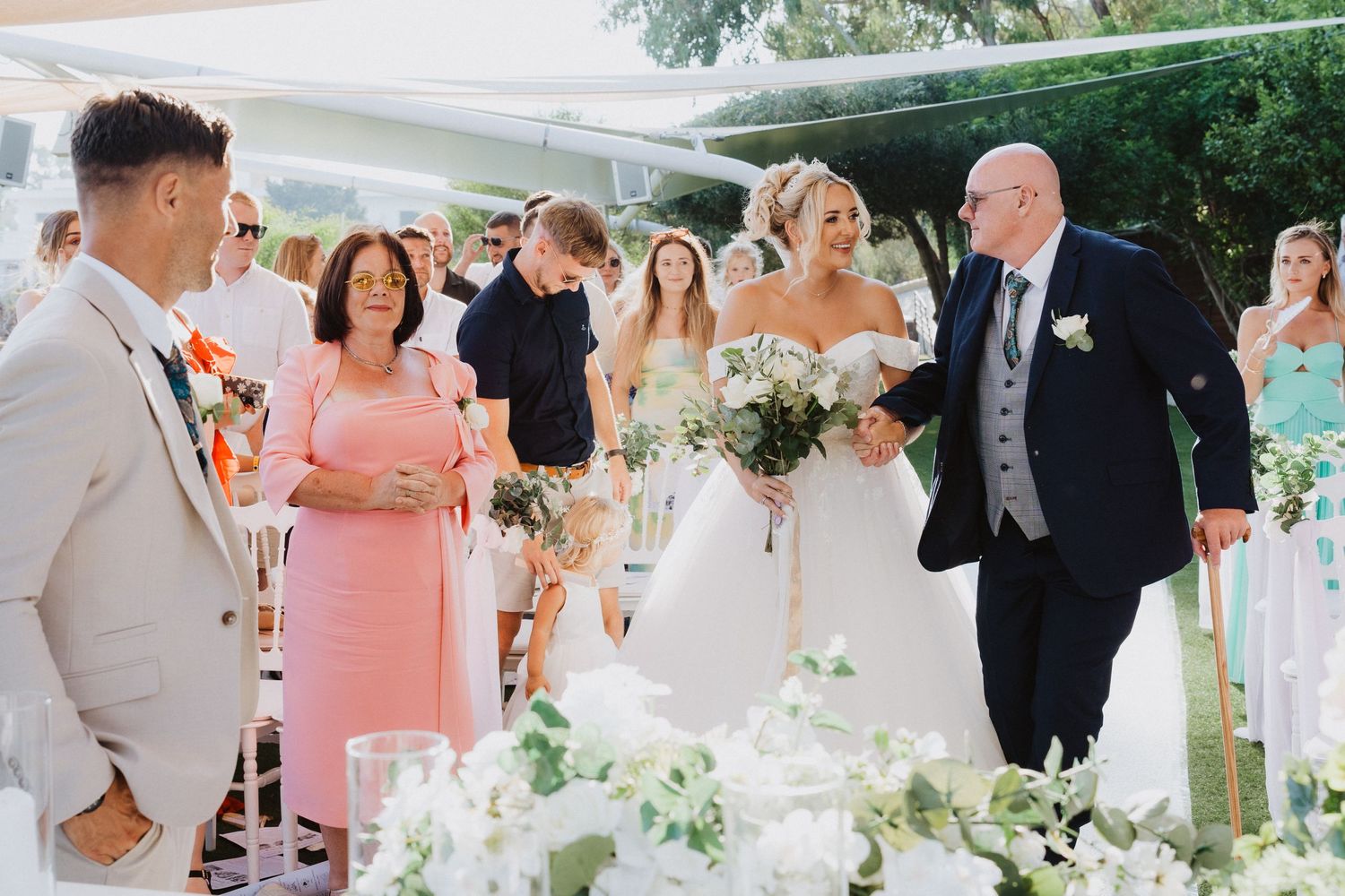 An outdoor wedding procession with guests in formal attire walking down a decorated aisle.