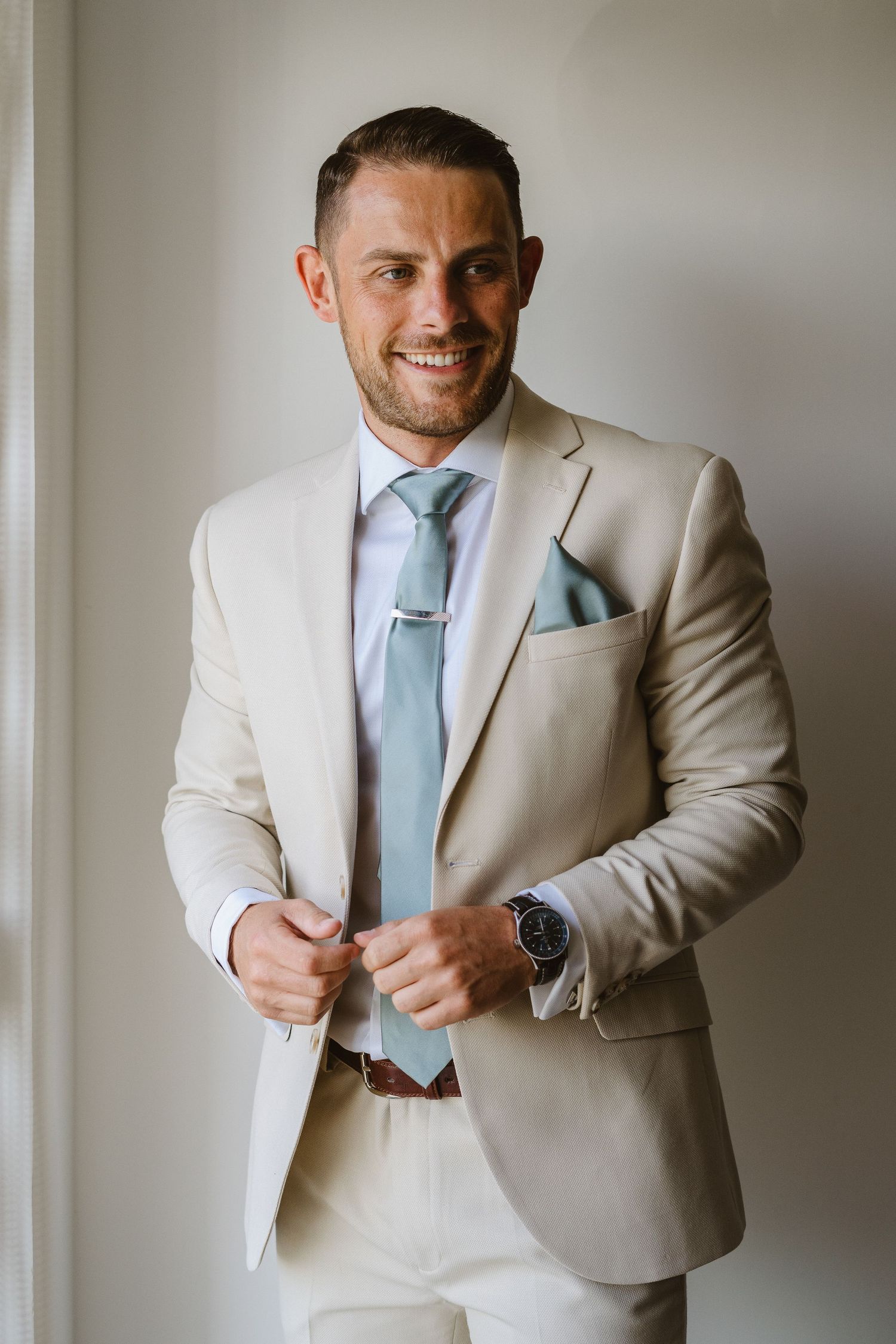 Elegant groom in beige suit and powder blue tie smiles while getting ready.