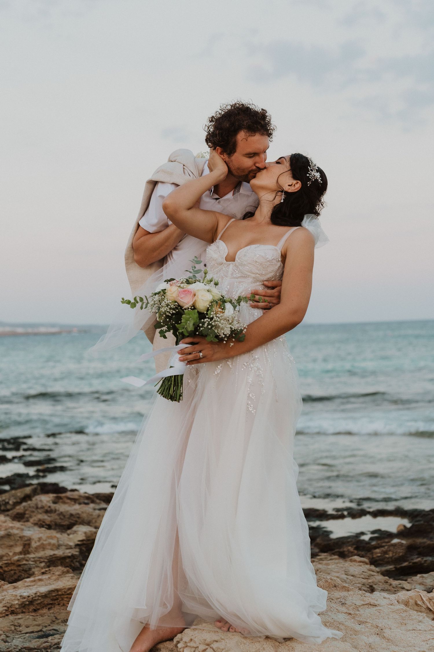 Intimate beachside wedding moment captured with couple embracing on coastal rocks at sunset.