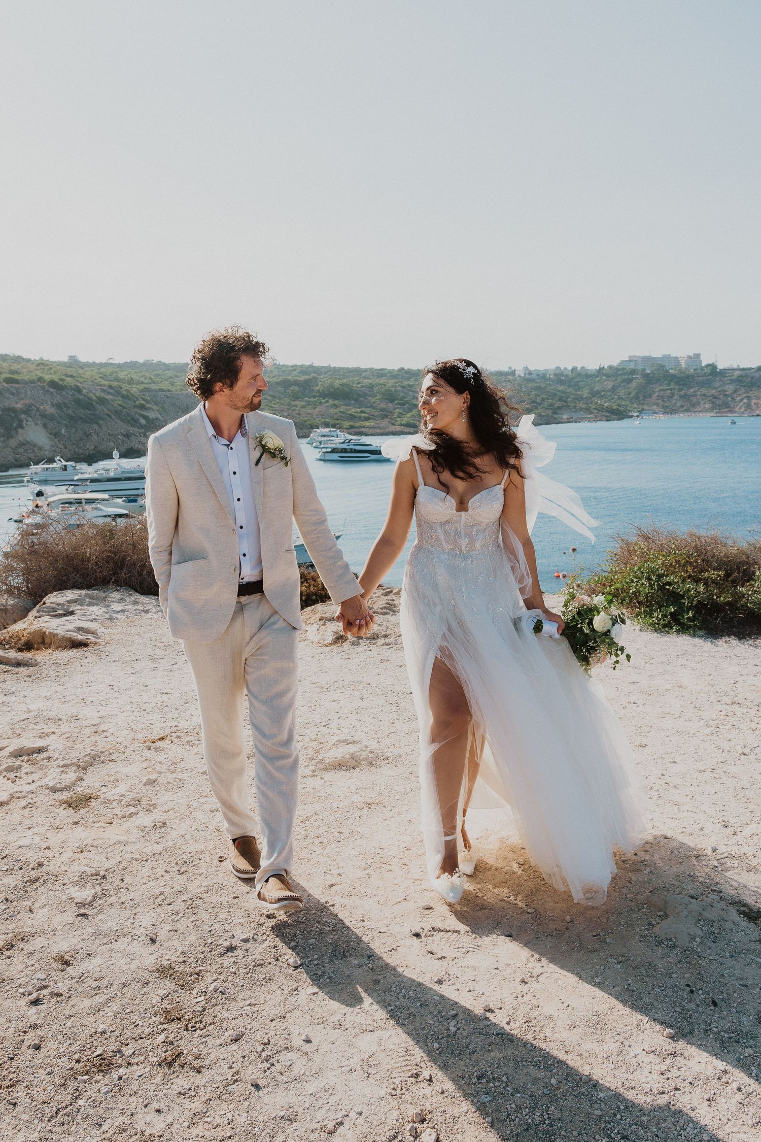 A wedding couple walks hand in hand along a coastal path with boats visible in the background.