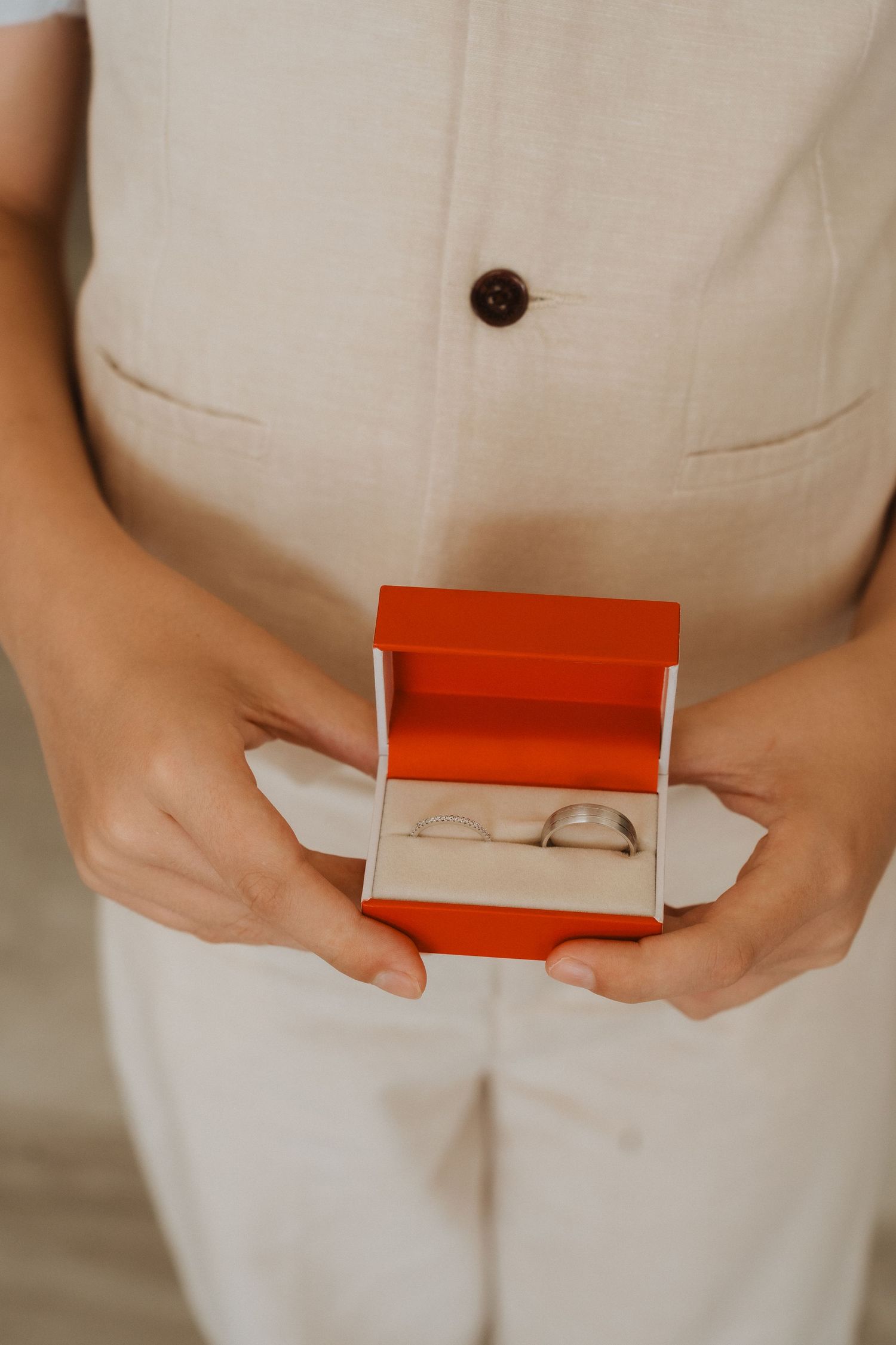 Hands holding an open red ring box with a silver ring nestled on white fabric against a cream-colored outfit.