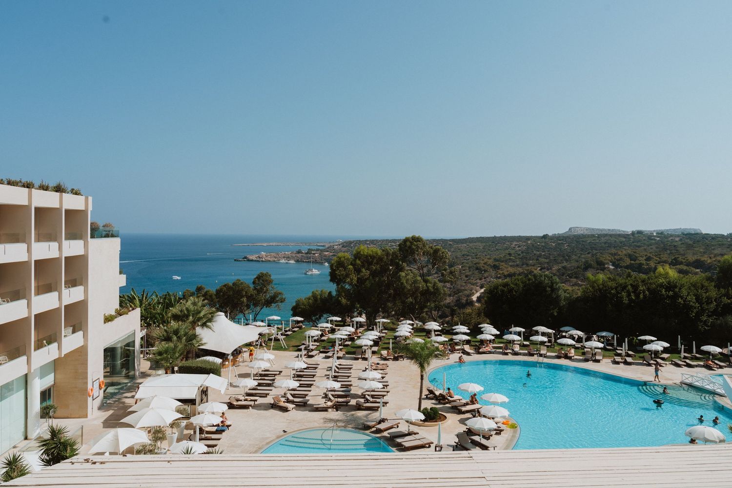 Luxury seaside resort with curved swimming pool overlooking turquoise Mediterranean waters and manicured grounds.
