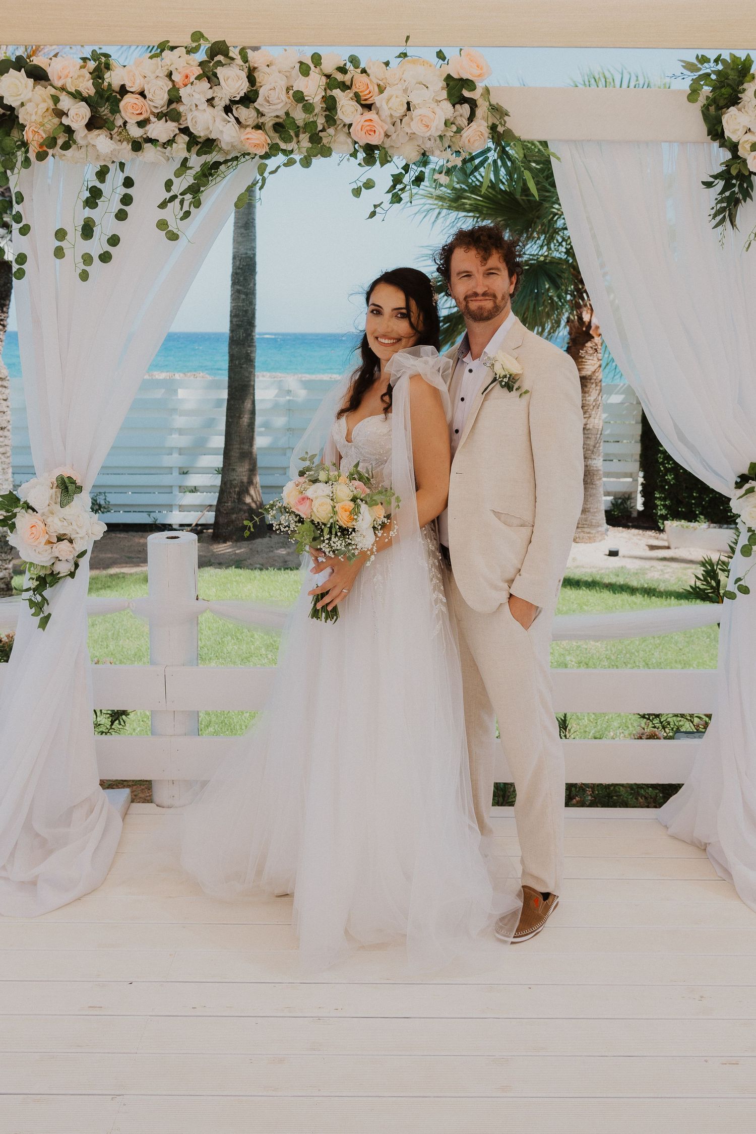 Wedding couple poses under floral arch with ocean view and white drapes.