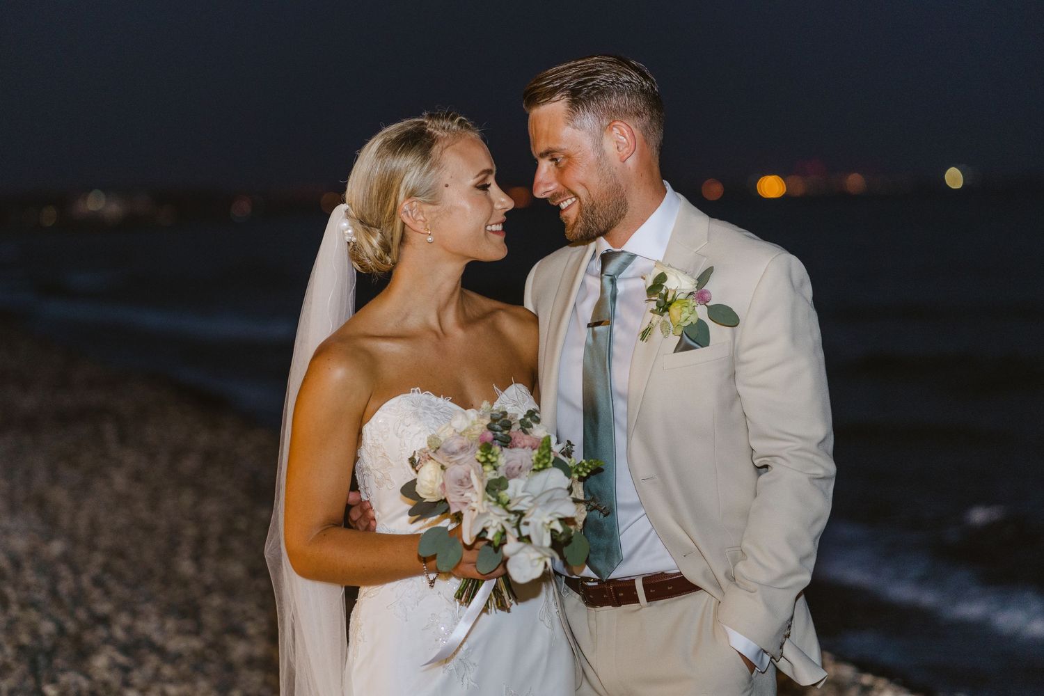 A beach wedding couple gazes at each other lovingly while holding a white floral bouquet at dusk.