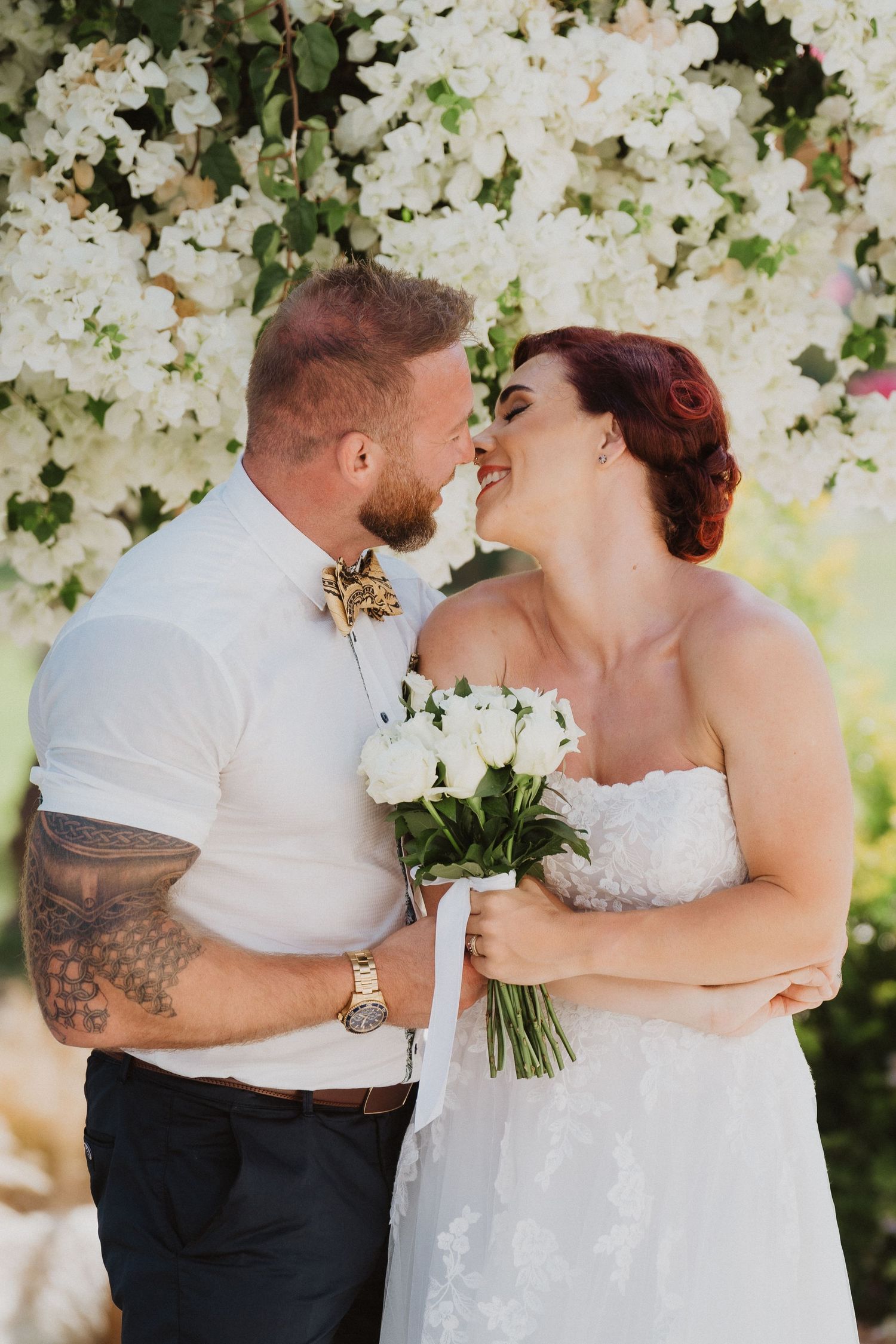 A romantic wedding moment captured under blooming white flowers on a sunny day.