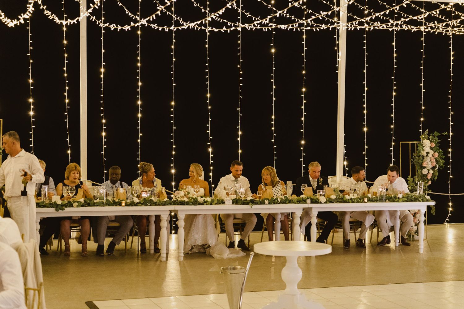 Wedding reception dinner setup with head table under cascading string lights.