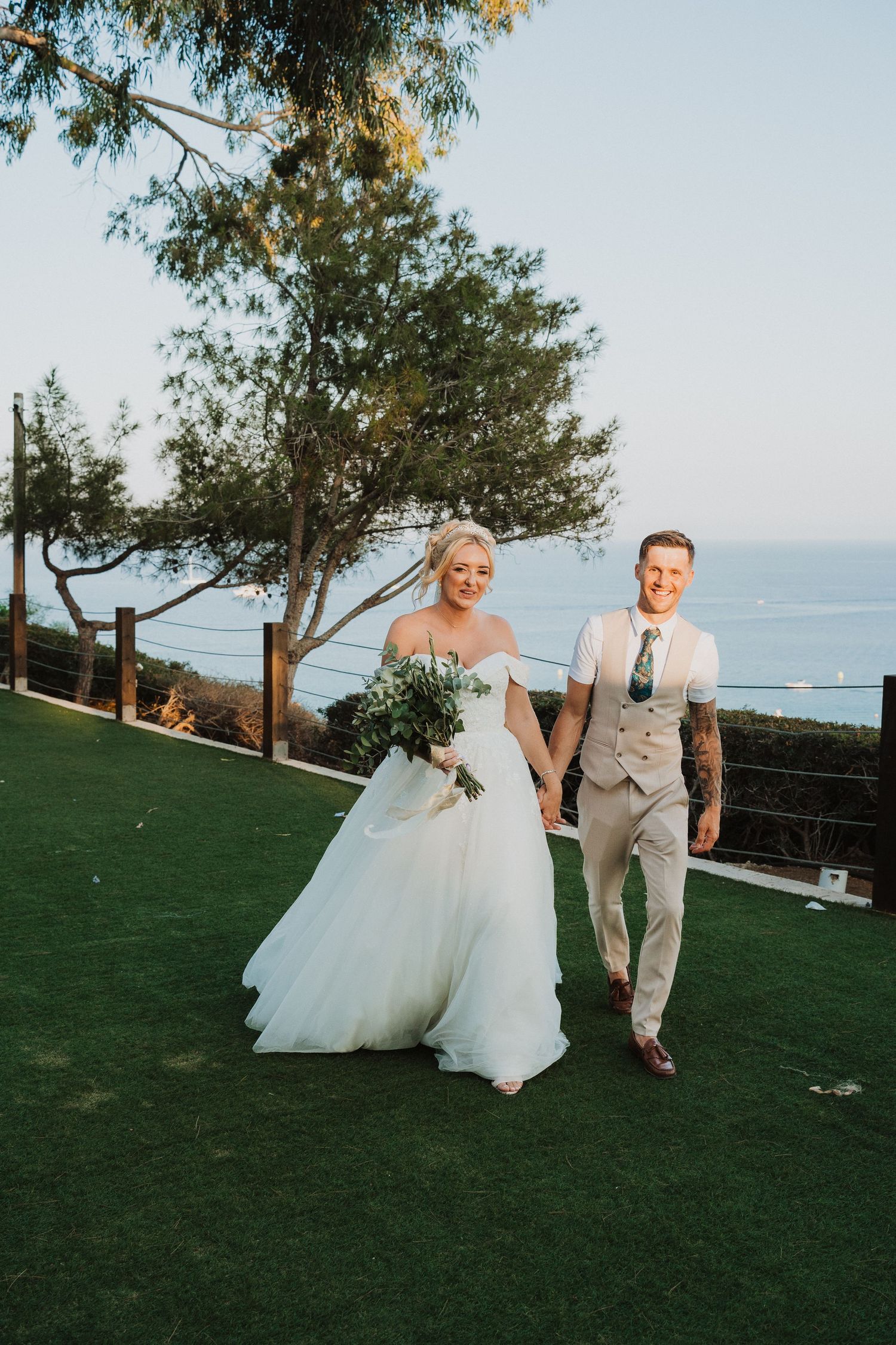 Newlyweds hold hands while walking on grass with coastal views and trees behind them.