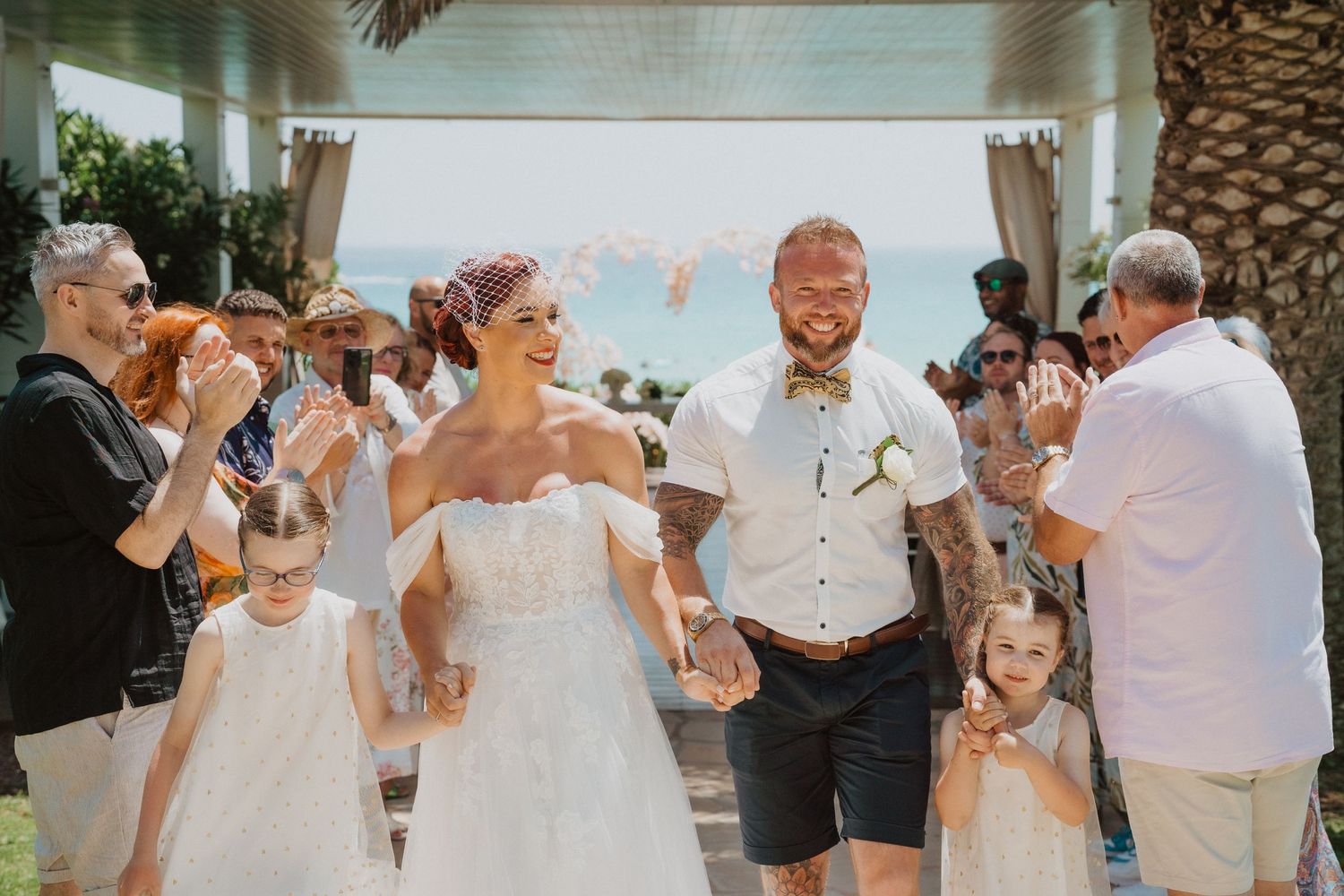 Wedding party exits ceremony under palm trees with guests applauding.