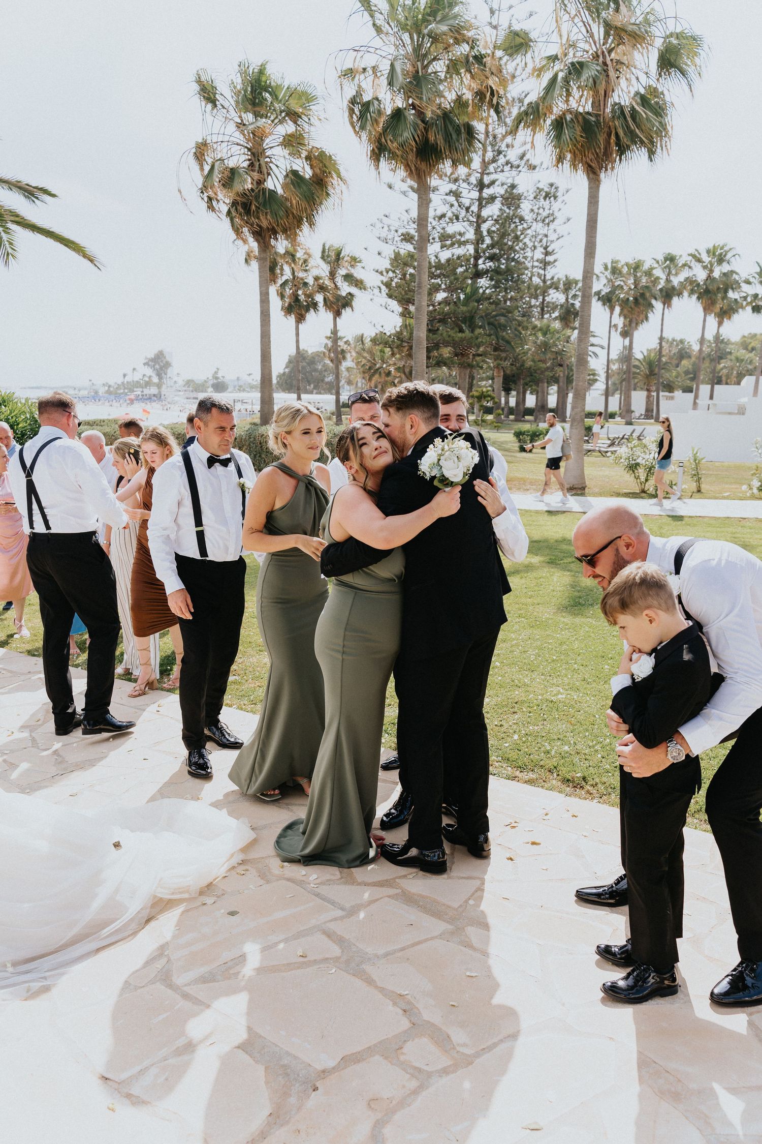 Wedding party in sage green dresses and black suits gather on stone patio surrounded by palm trees.
