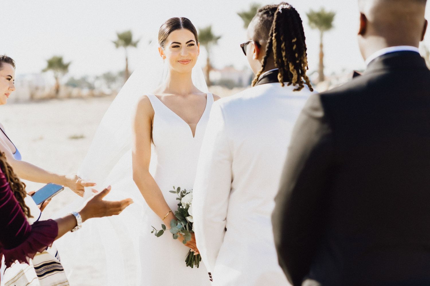 Outdoor beach wedding ceremony with white floral arch and guests looking on.