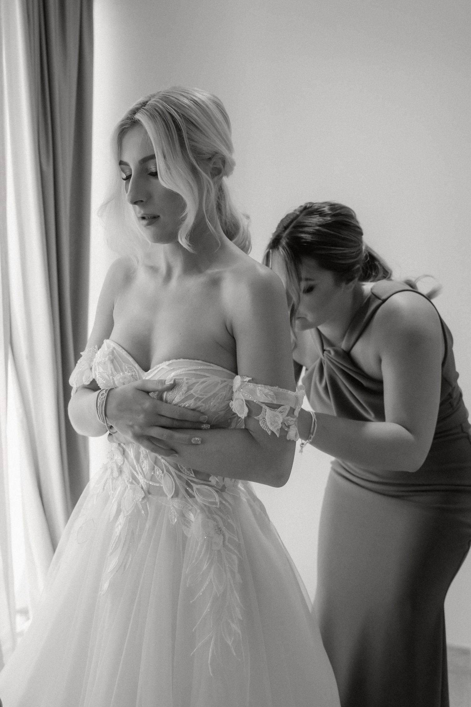 A tender moment during bridal preparations as a bridesmaid helps adjust the wedding dress.