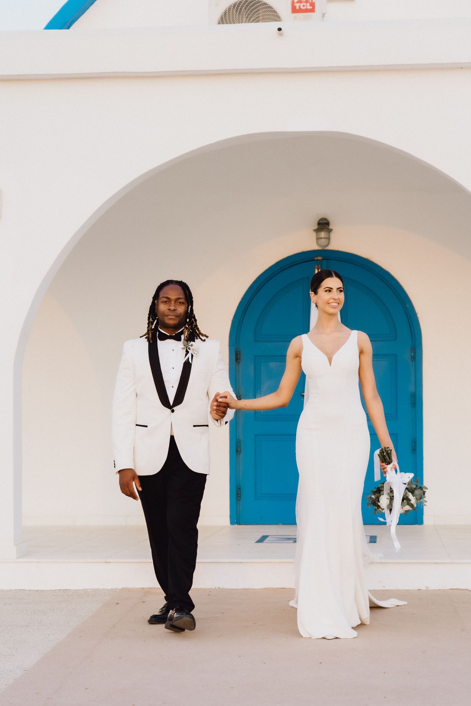 Elegant couple in white formal attire stands before a bright blue door.