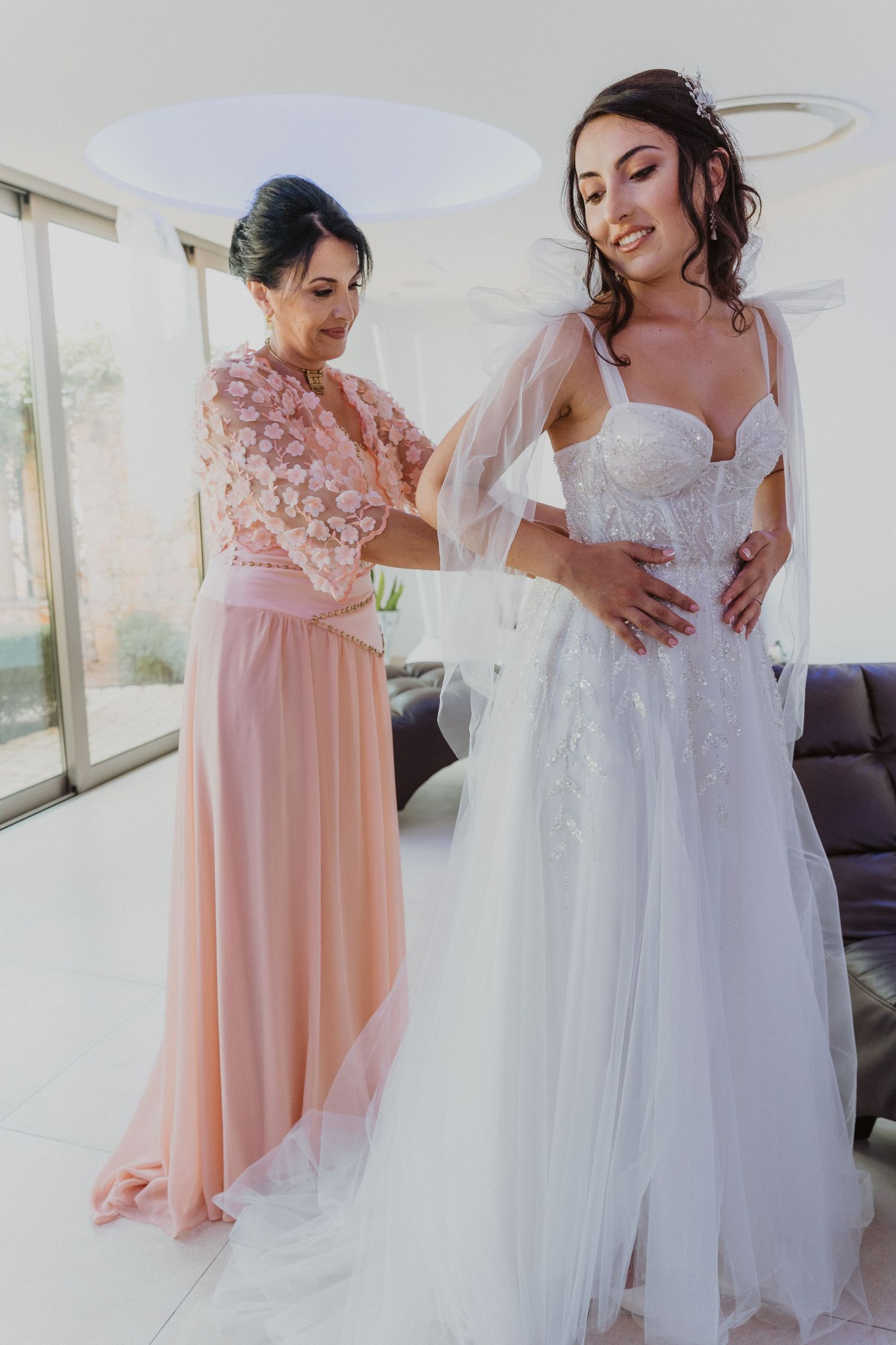 Bridesmaid in pink helps bride with final adjustments to her elegant white wedding gown.
