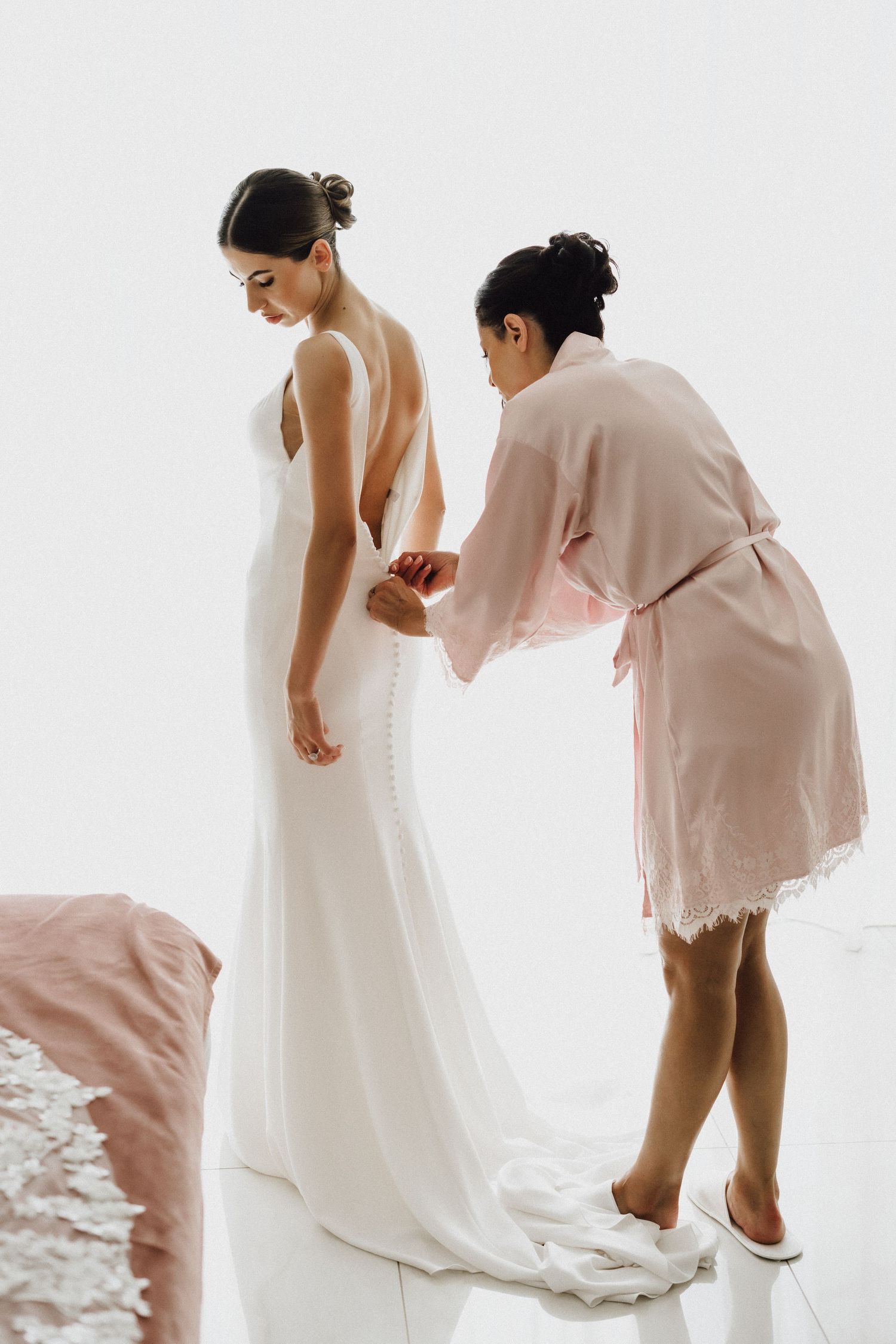 Elegant white wedding dress being adjusted from behind by person in pink robe.
