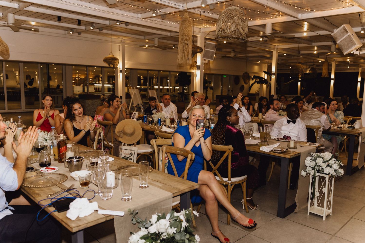 Indoor reception space filled with wedding guests dining at decorated tables under industrial ceiling.