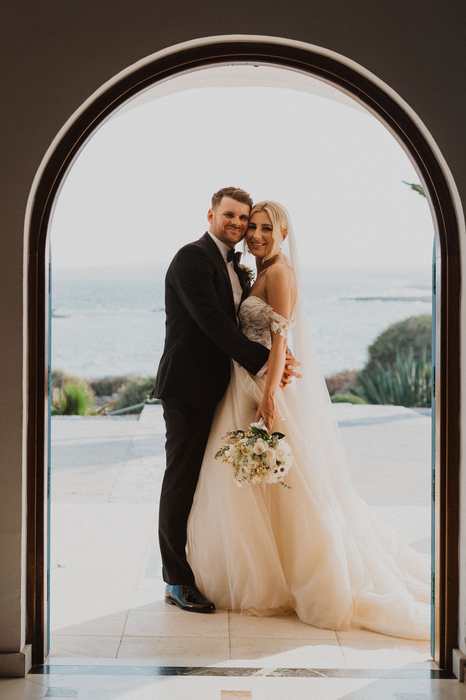 Joyful wedding portrait framed by arched doorway with seaside backdrop.